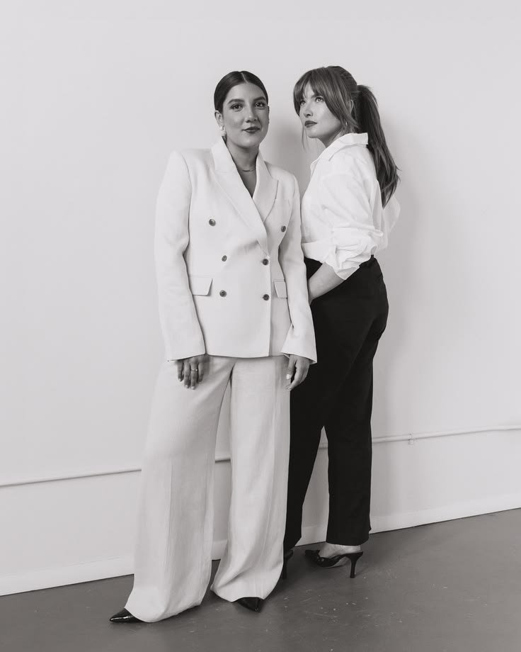 Two women standing against a plain wall, one in a white suit with wide-leg pants, and the other in a white blouse and black pants, both wearing high heels.