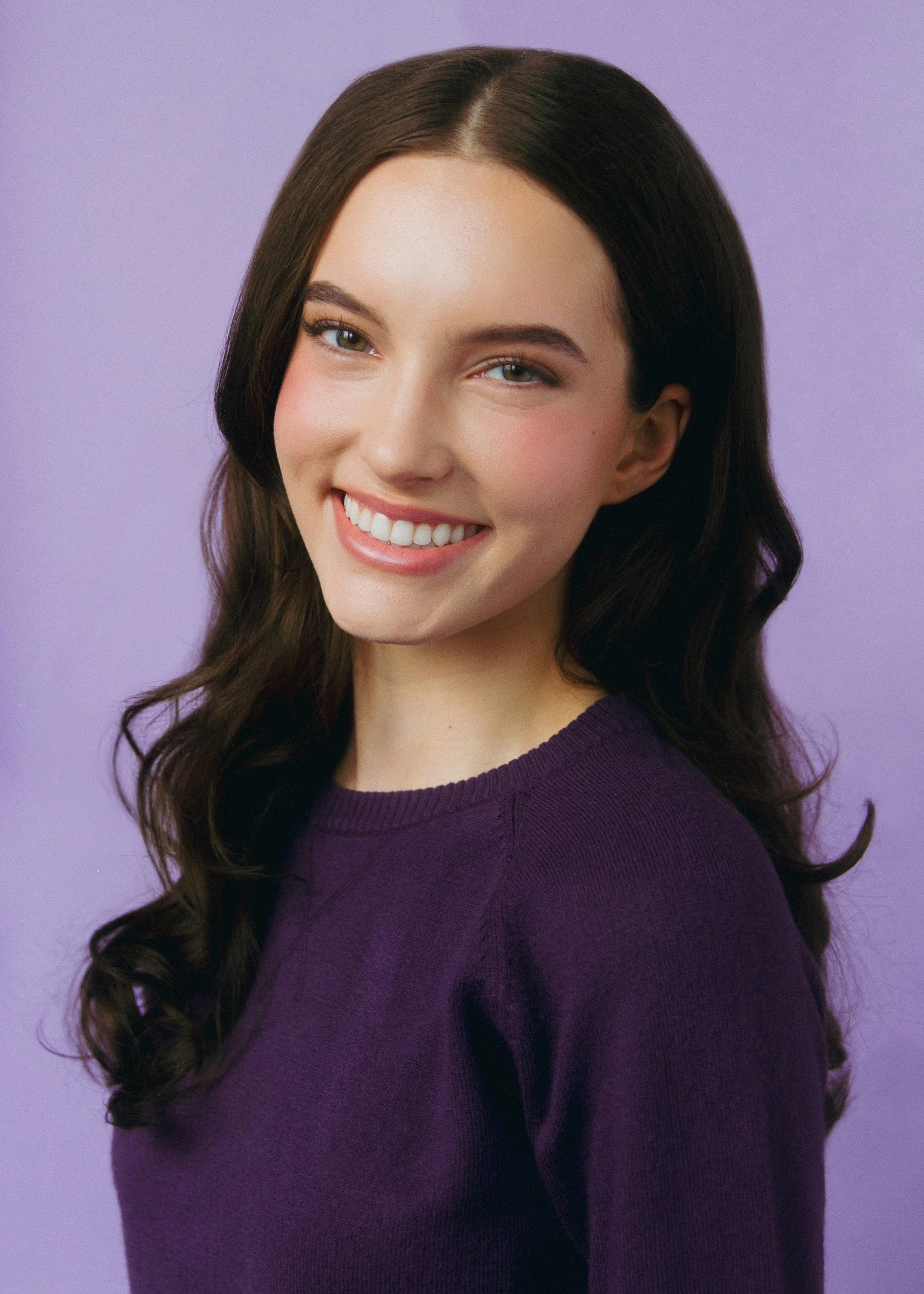 A young woman with long brown wavy hair wearing a purple sweater, smiling with white teeth, against a pastel purple background.