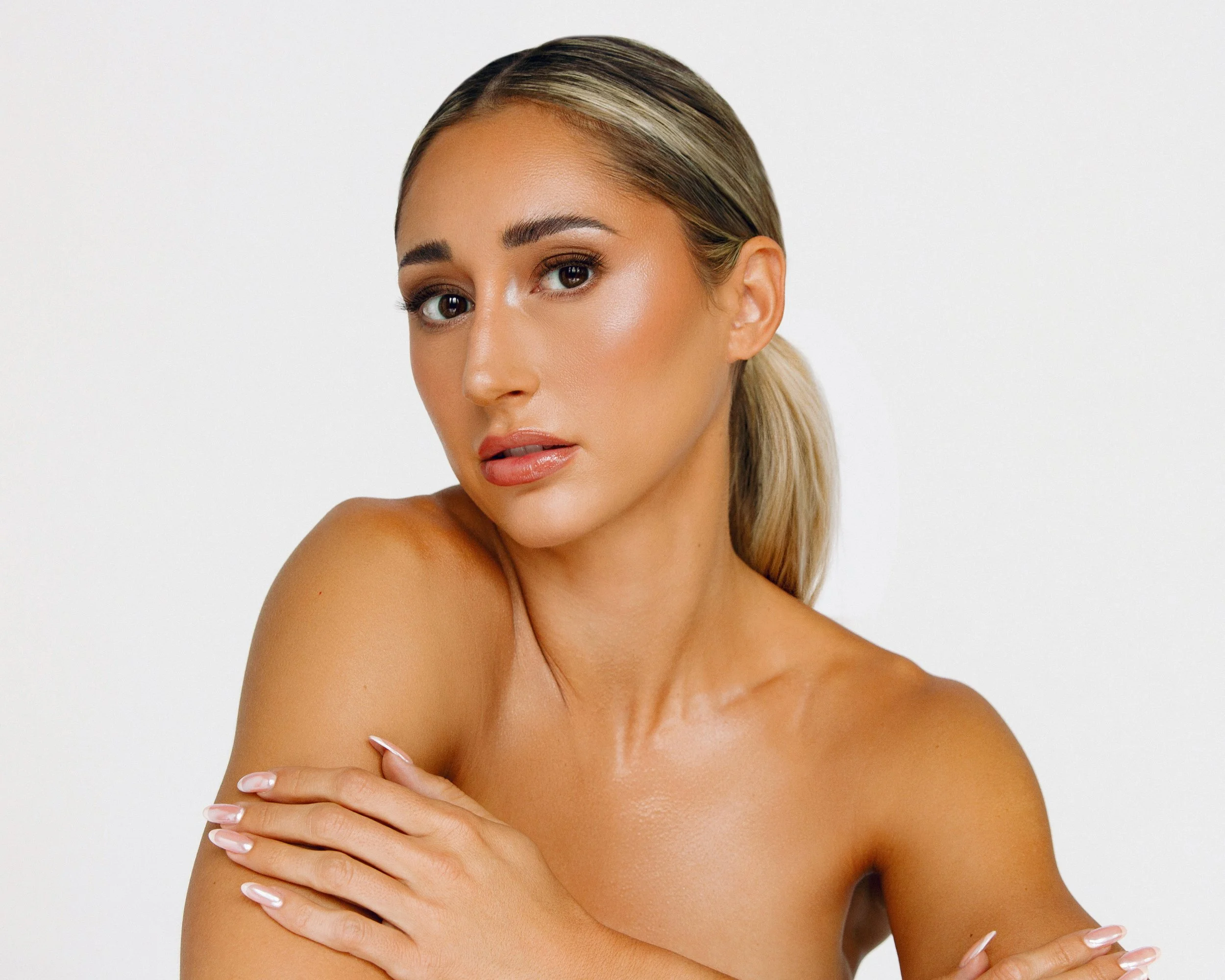 A woman with blonde hair and makeup posing against a plain white background, covering her chest with her arm.