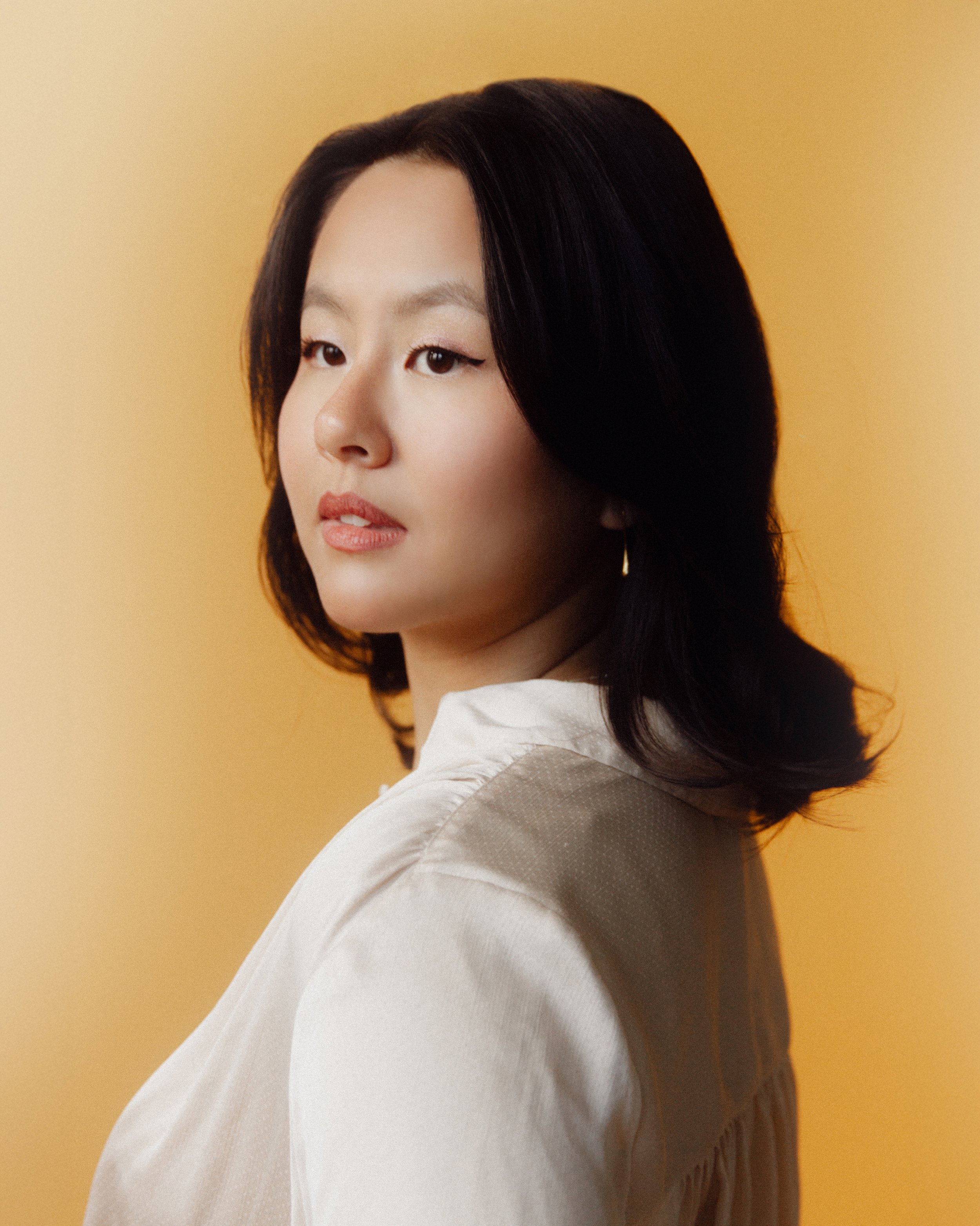 A young Asian woman with shoulder-length black hair gazes to her left against a yellow background, wearing a white top.