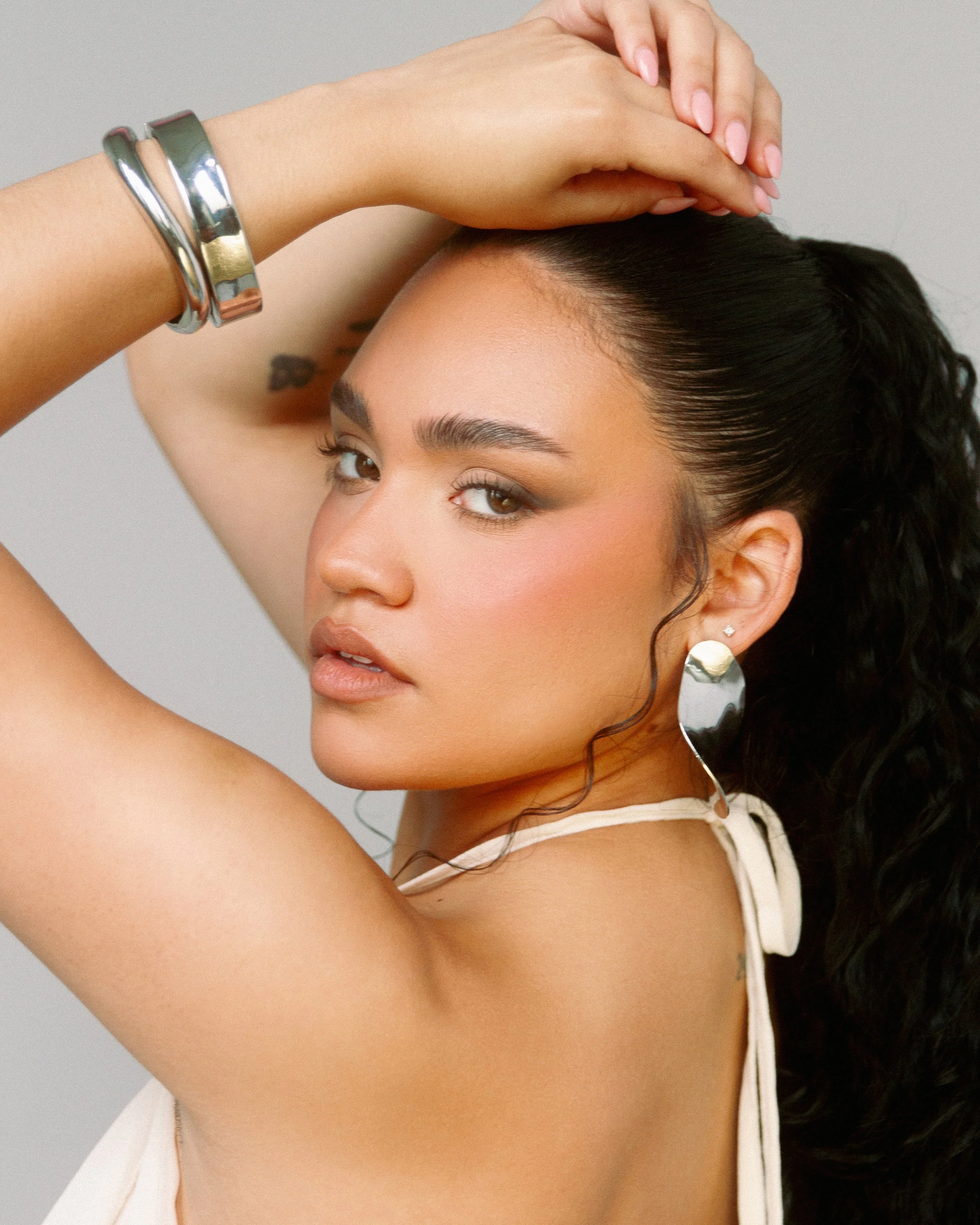 A woman with dark curly hair, wearing large shiny earrings and metallic bracelets, poses with one arm raised and her hand resting on her head. She has makeup on and is looking at the camera.