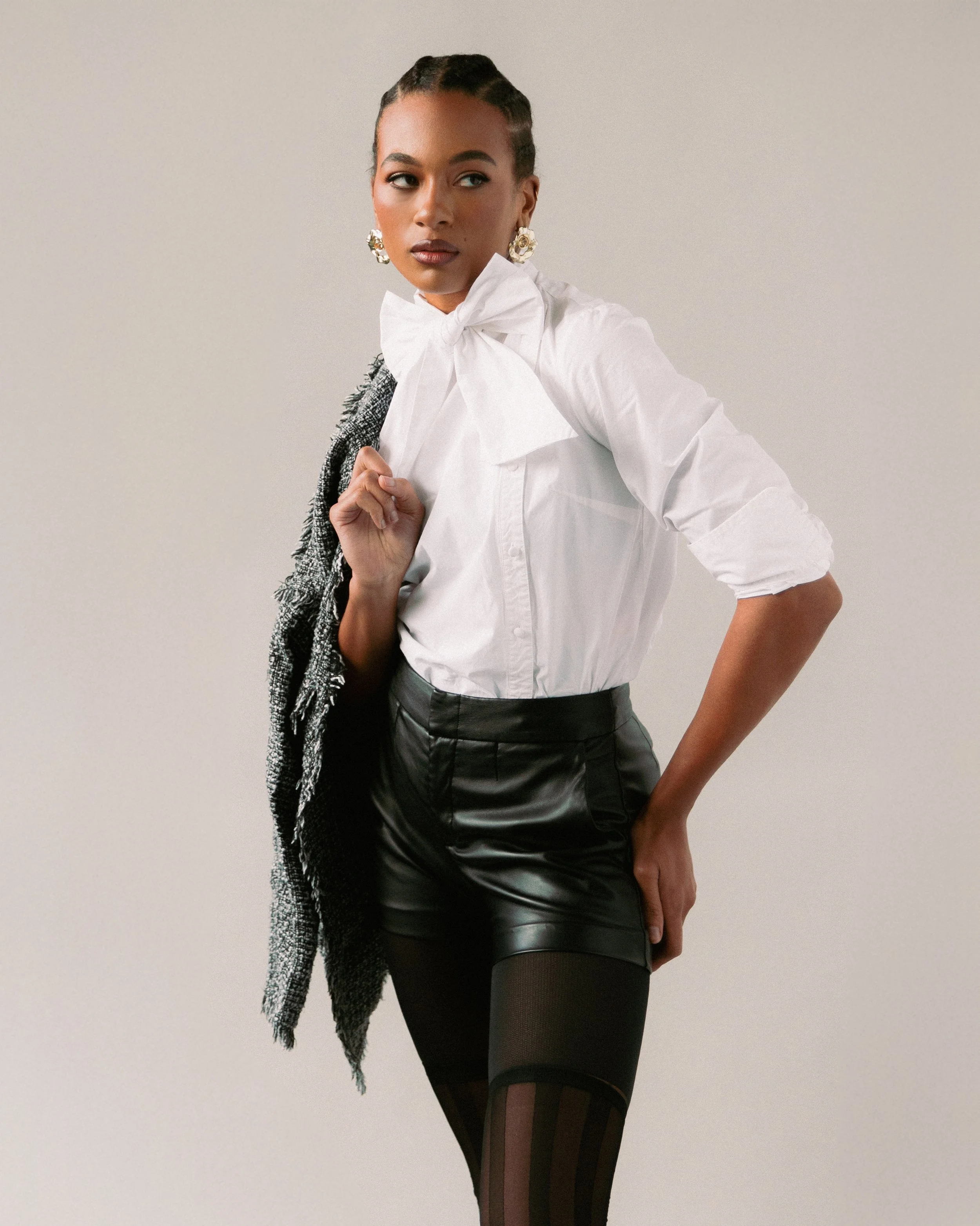 A woman wearing a white blouse with a large bow, black leather shorts, patterned tights, and gold earrings, standing against a plain background.