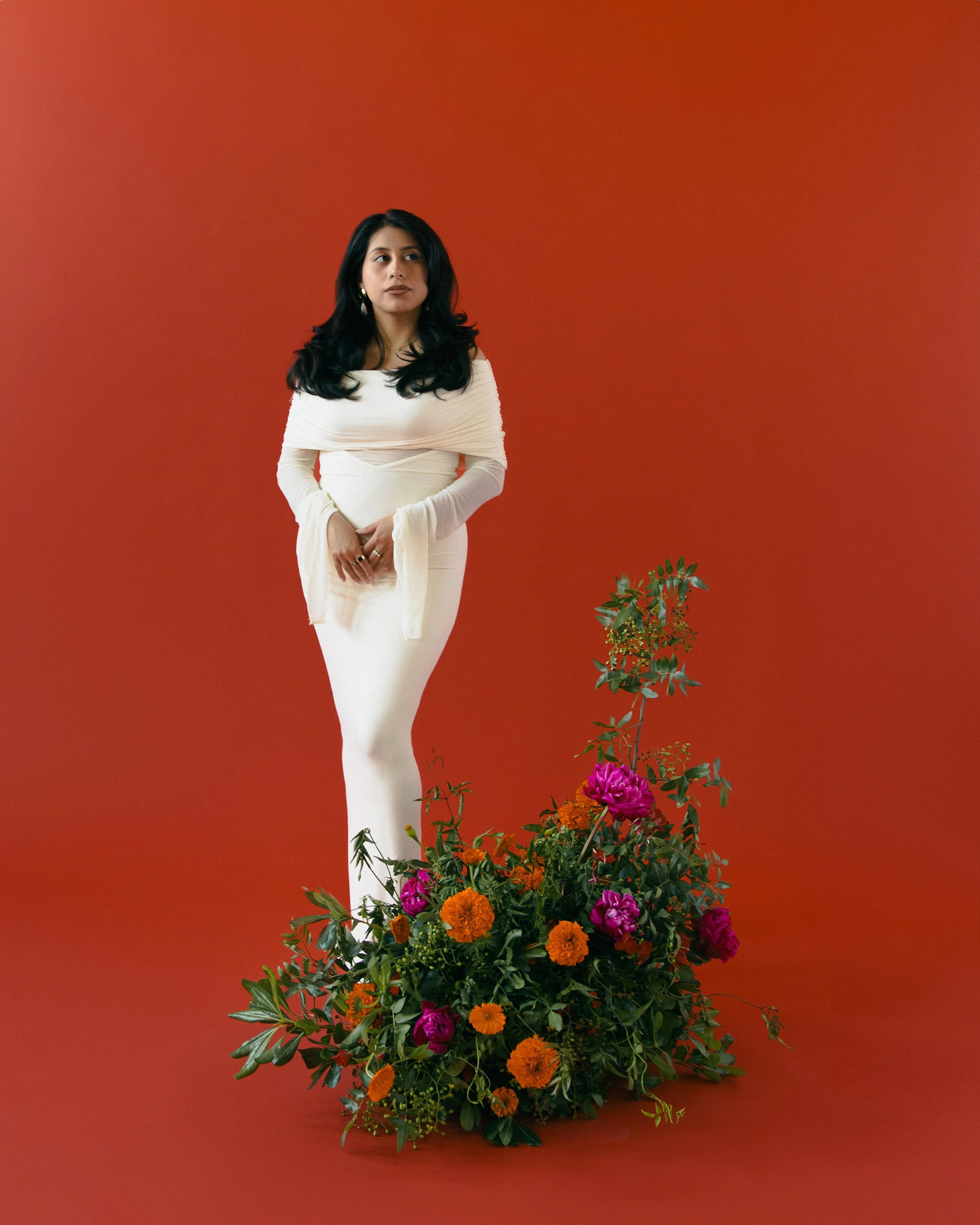 A woman in a white dress stands against a red background, with a large bouquet of colorful flowers including pink, orange, and red blooms and green foliage at her feet.