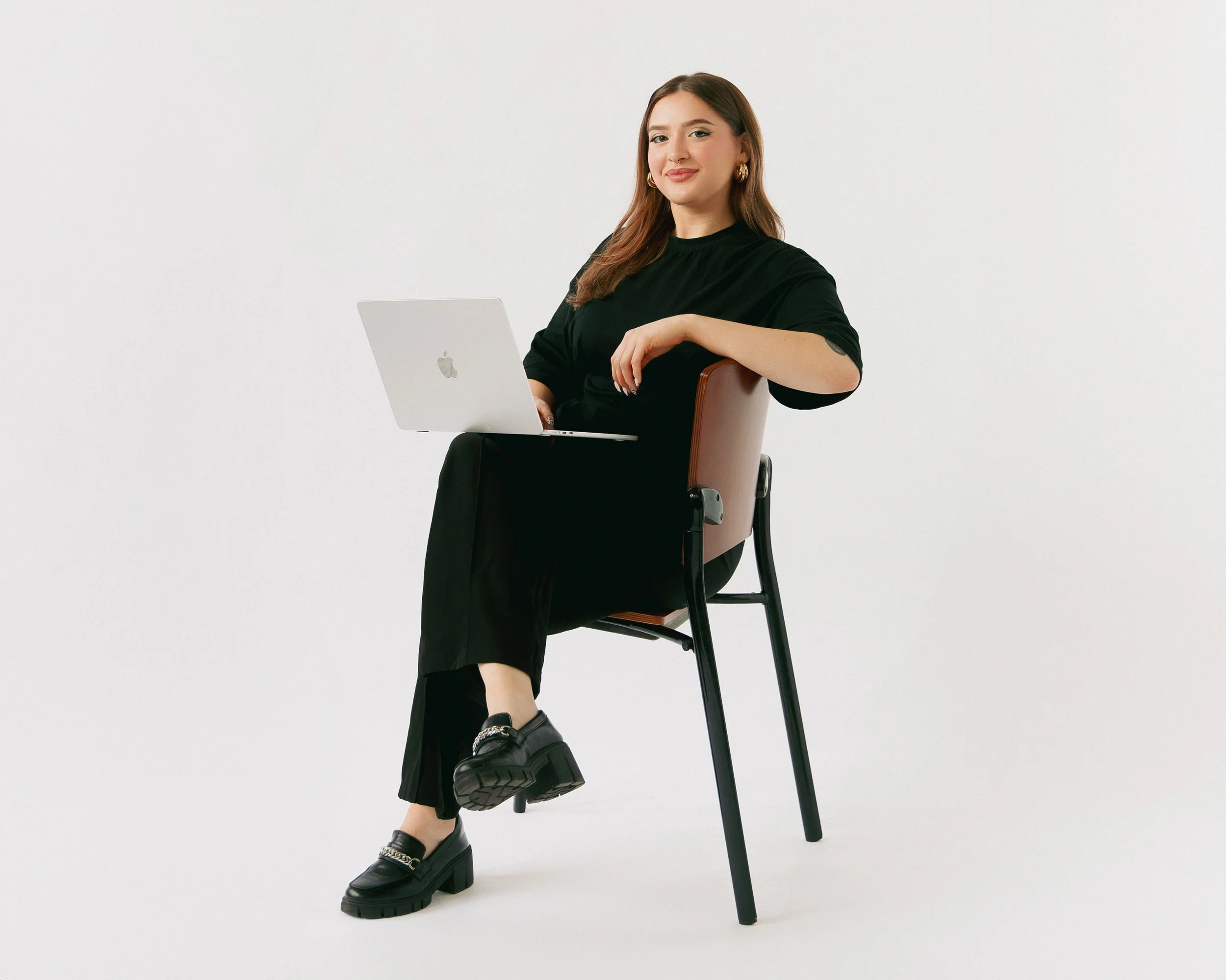 Woman sitting on a chair with one leg crossed over the other, wearing black clothing, black platform shoes, gold hoop earrings, and holding a laptop, against a plain white background.