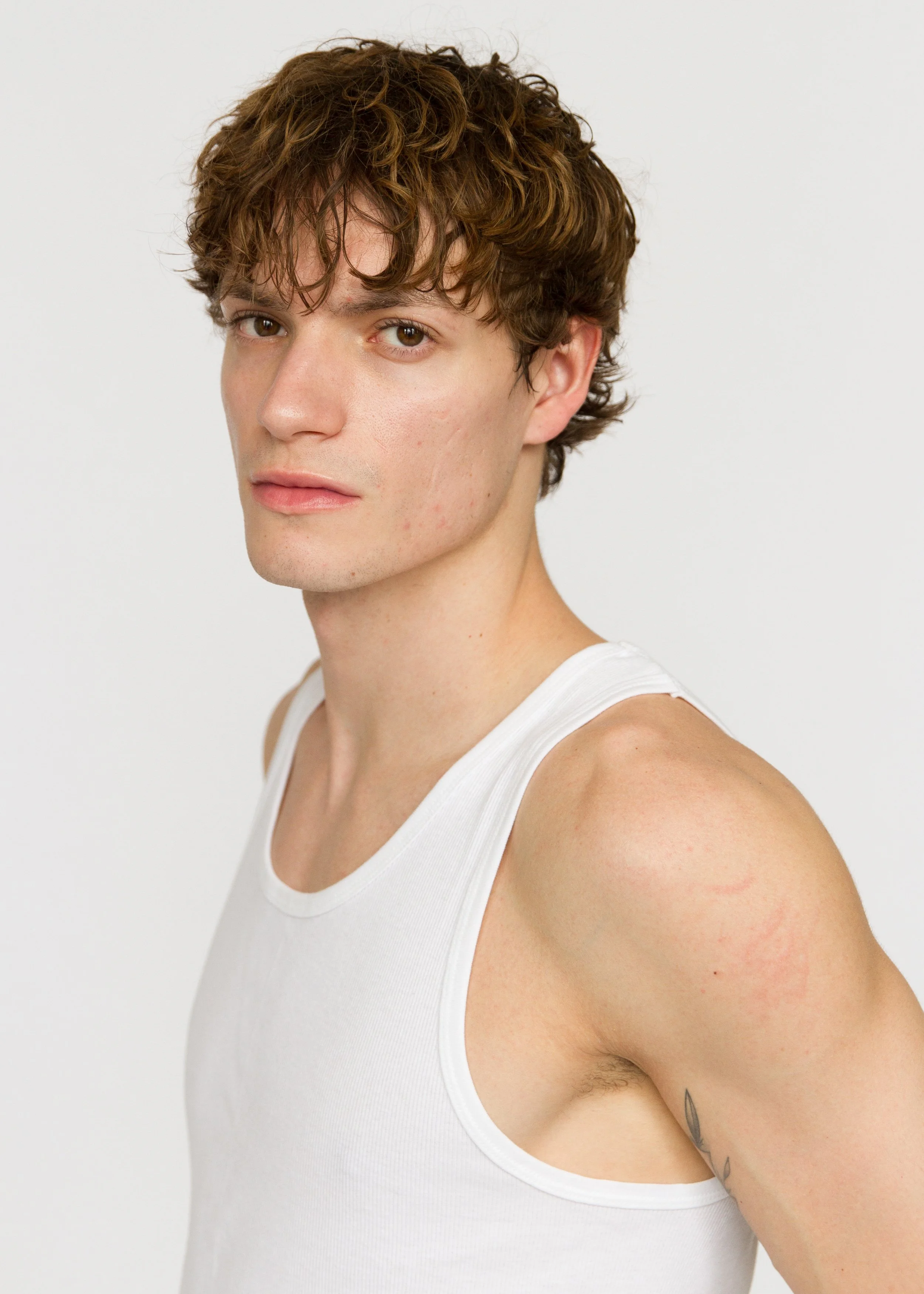 Close-up of a young man wearing a white tank top, with wavy brown hair, light skin, and a tattoo on his upper arm, standing against a plain white background. Model Digitals