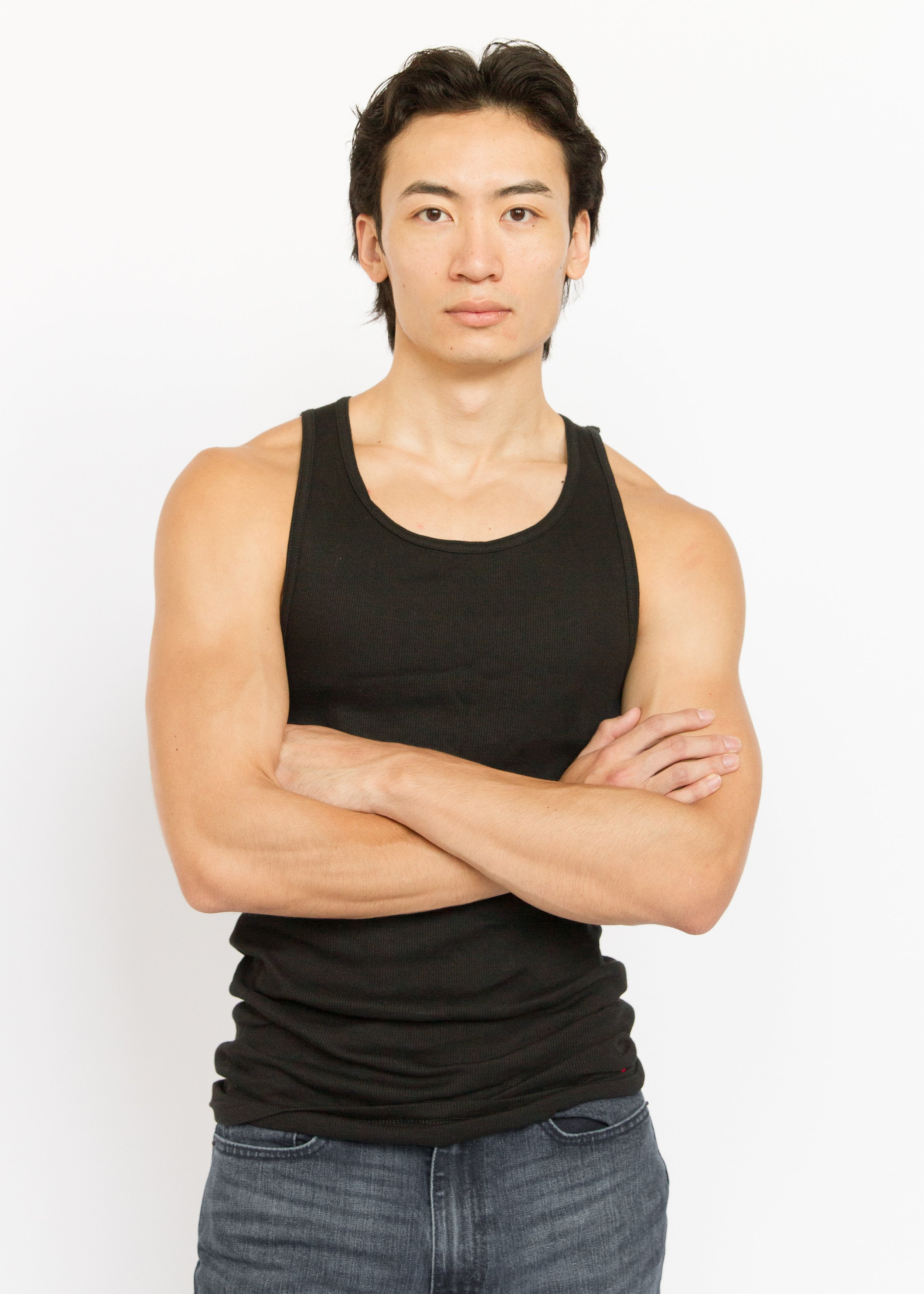 A young man with black hair wearing a black sleeveless shirt and dark jeans, standing with arms crossed against a plain white background.