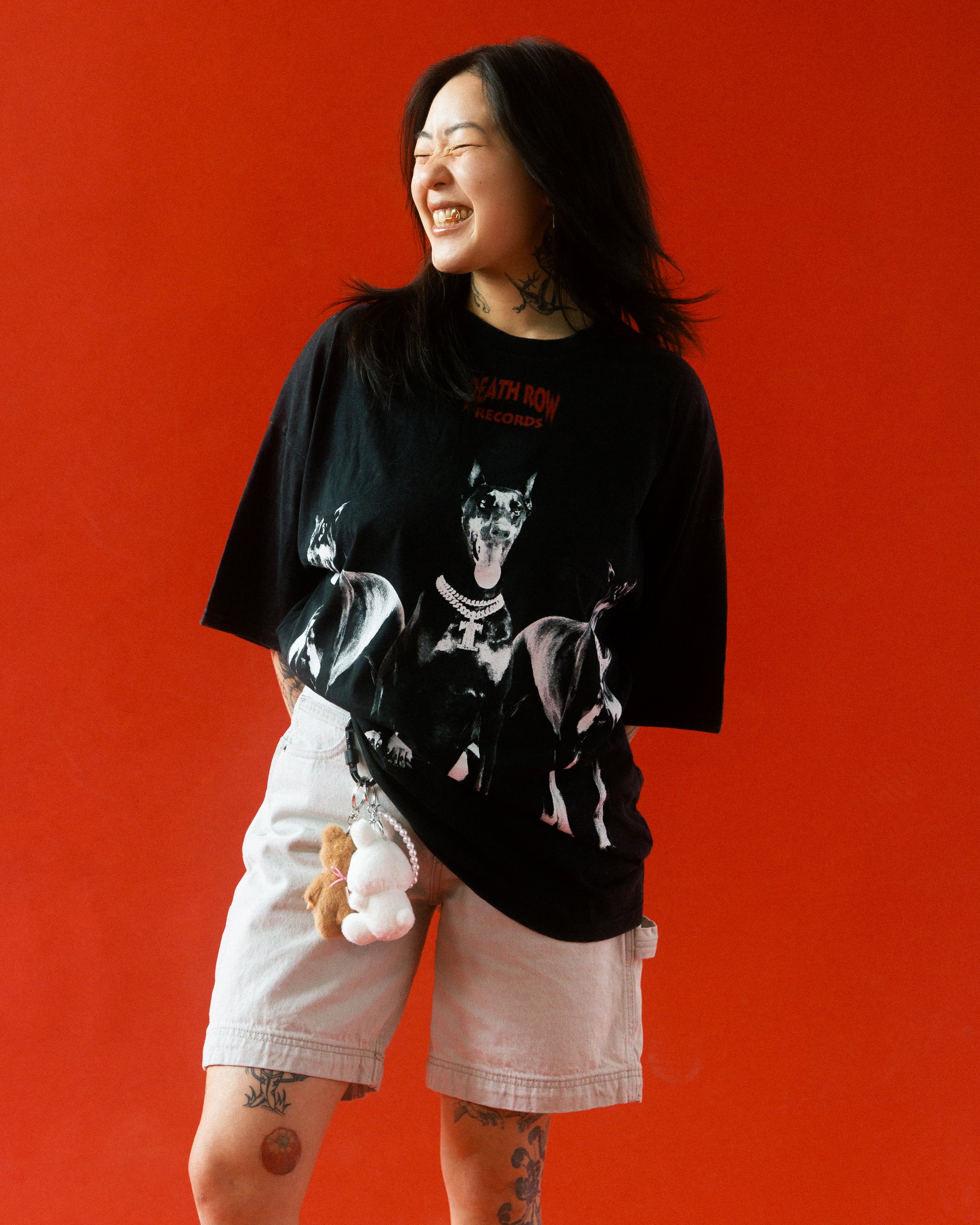 A woman with black hair, tattoos, and a big smile standing against a red background, wearing a black graphic t-shirt and light-colored shorts with plush toy keychains hanging from her pocket.