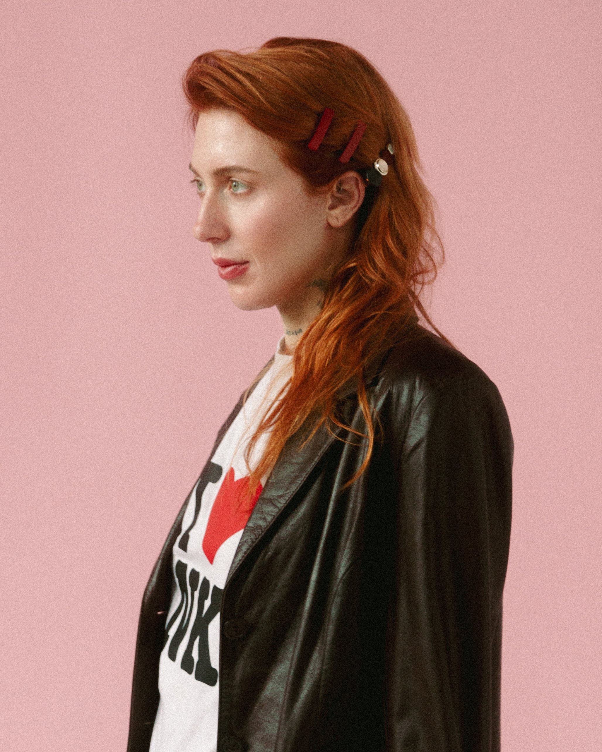 A woman with long red hair wearing a black leather jacket and a white T-shirt with a graphic design, standing against a pink background.