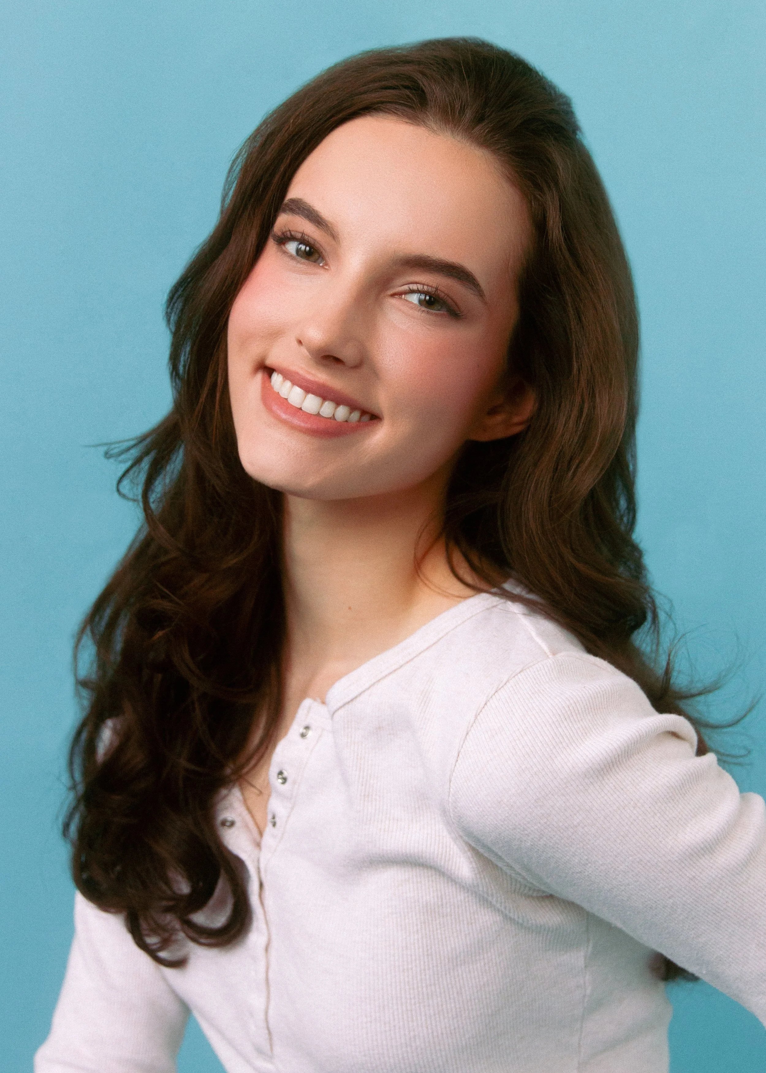 A young woman with long, wavy brown hair smiling against a light blue background.