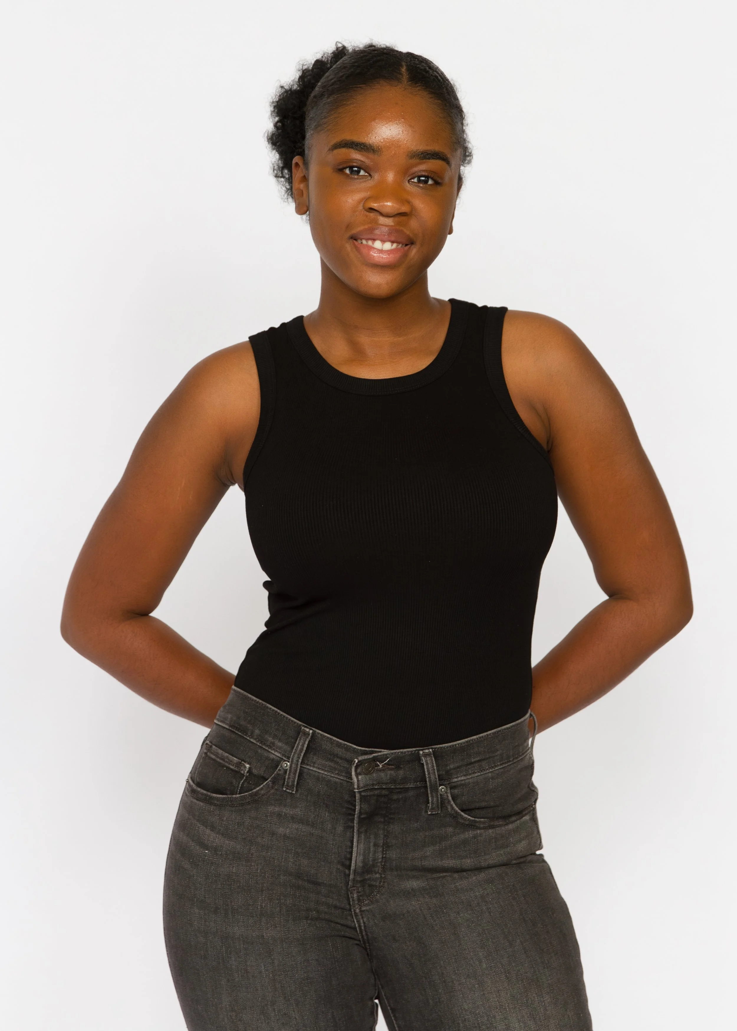 A woman with dark skin, curly hair styled in a puff, wearing a black sleeveless top and dark jeans, standing against a plain white background, smiling with her hands on her hips.