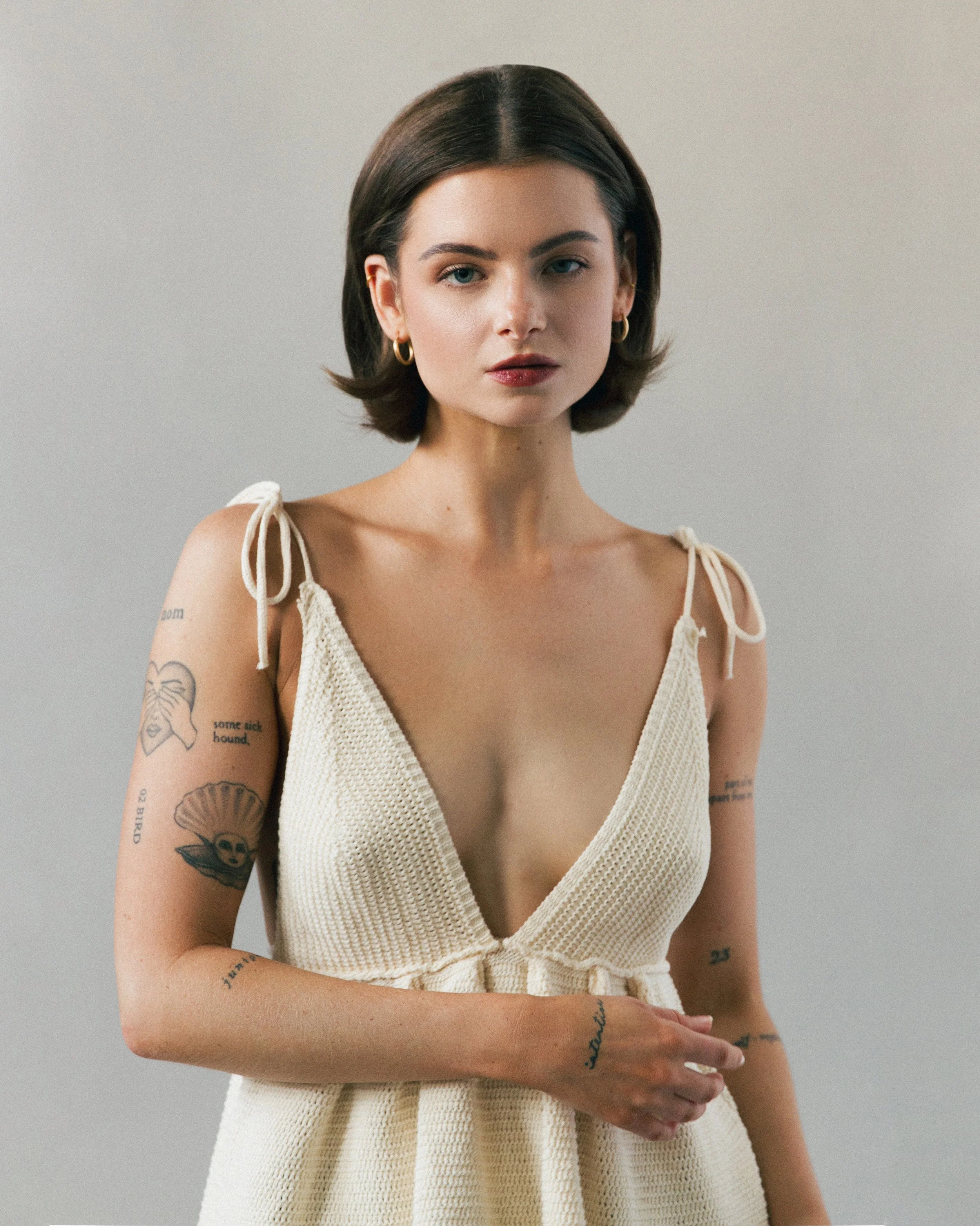 A young woman with dark brown hair, blue eyes, and light skin, wearing a cream-colored knitted top with tie straps, gold hoop earrings, and visible tattoos on her arms, stands against a plain light gray background.