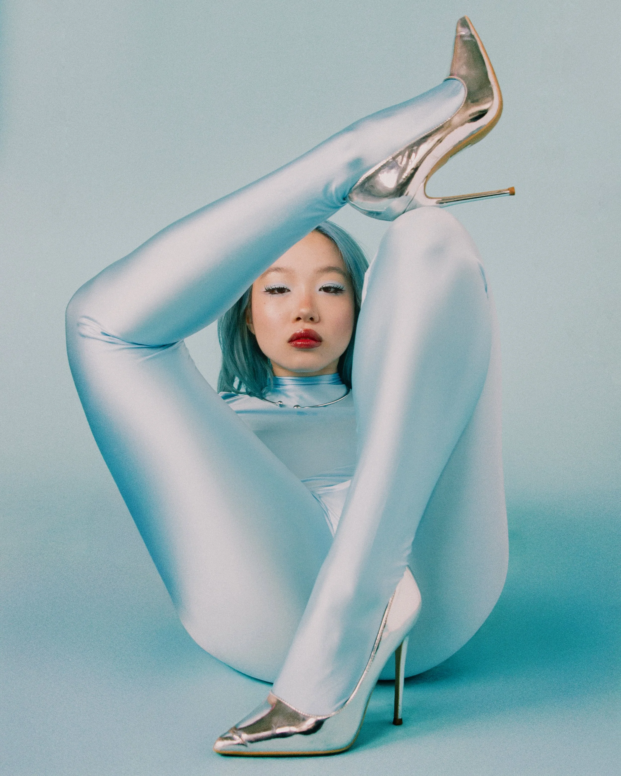A woman with blue hair in a shiny, tight-fitting silver bodysuit and matching high heels posed with her legs up and crossed in front of her, against a light blue background.
