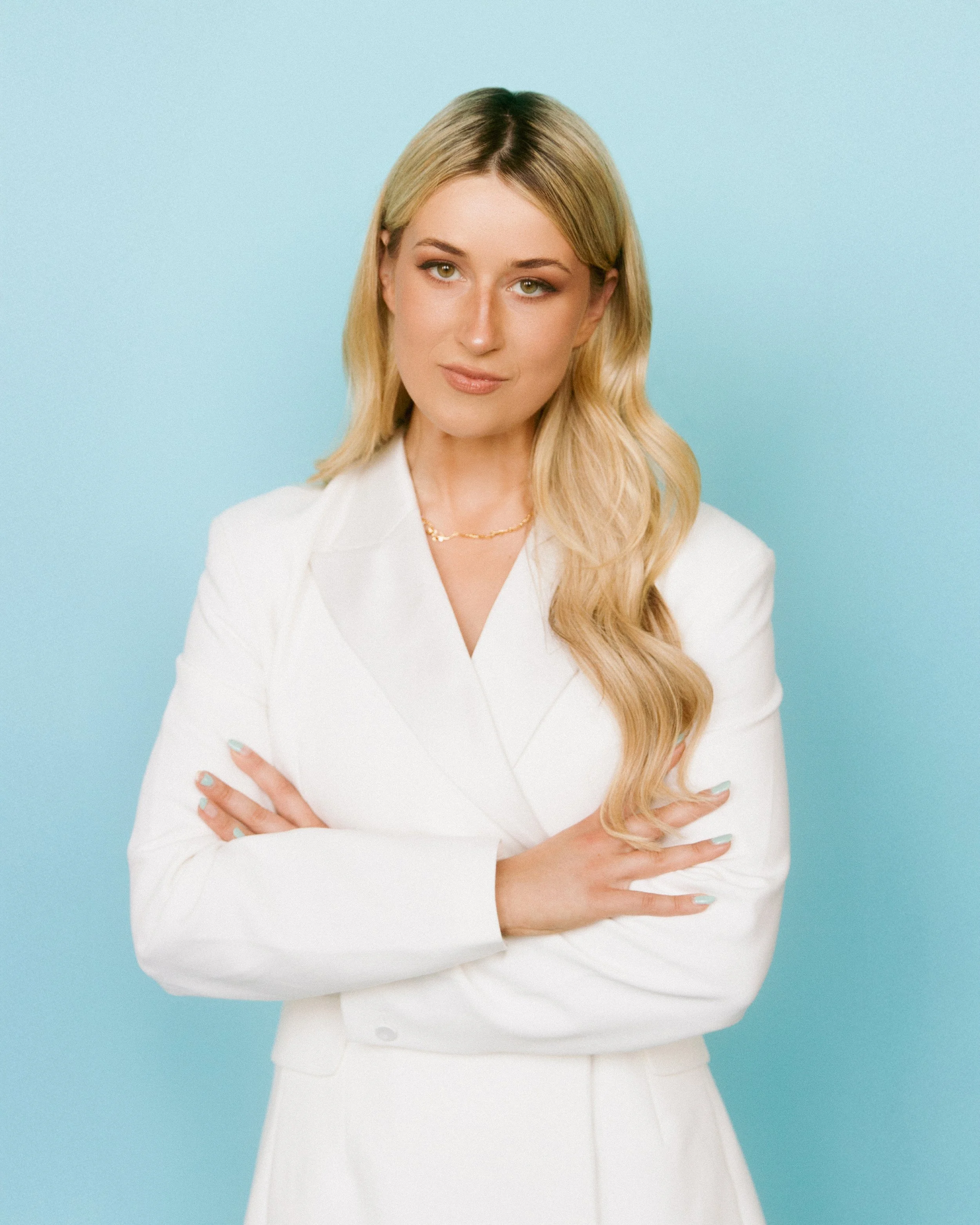 A woman with long blonde hair wearing a white blazer and gold necklace poses with her arms crossed in front of a light blue background.