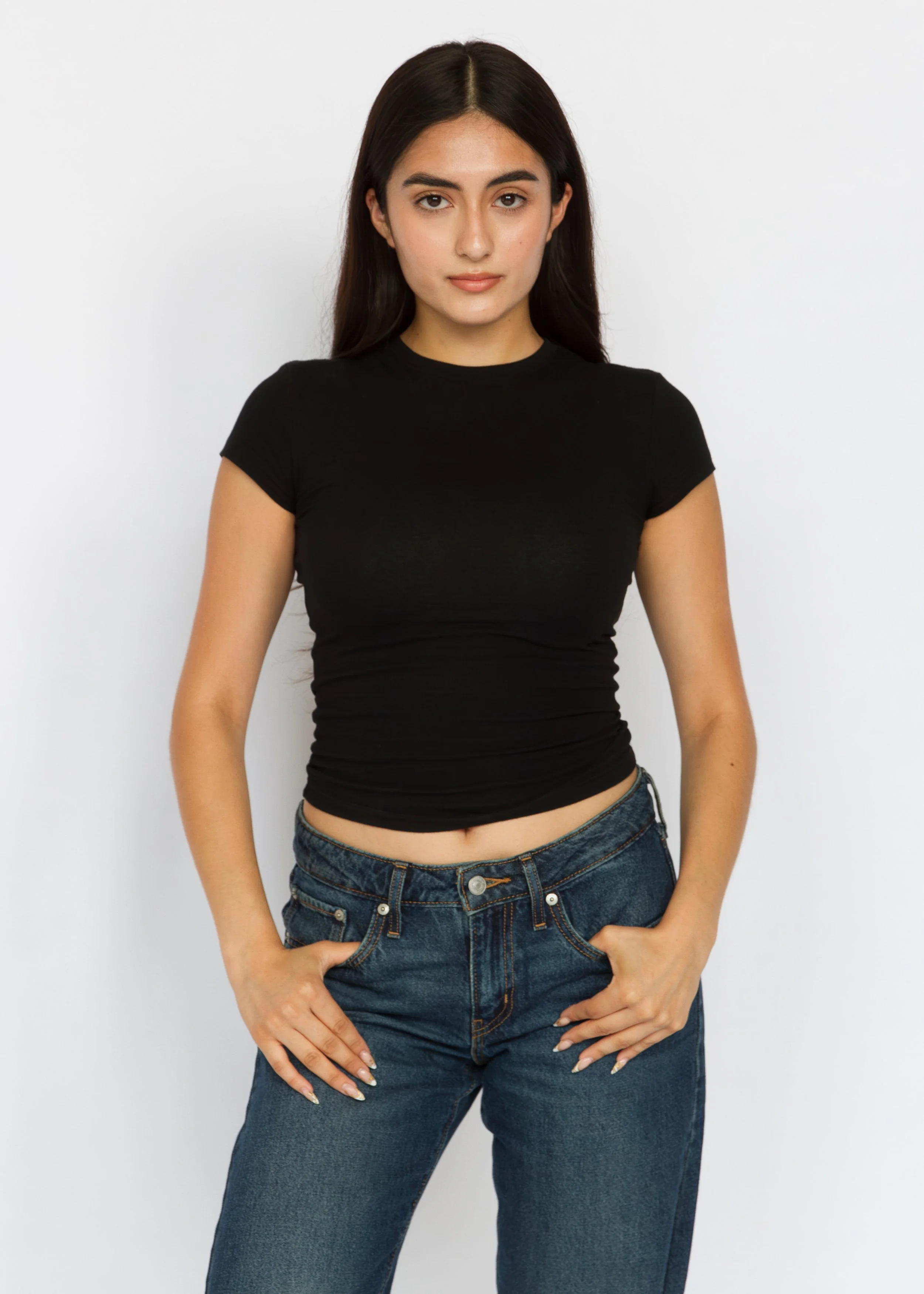 Young woman with long dark hair wearing a black t-shirt and blue jeans against a plain white background. Model Digitals