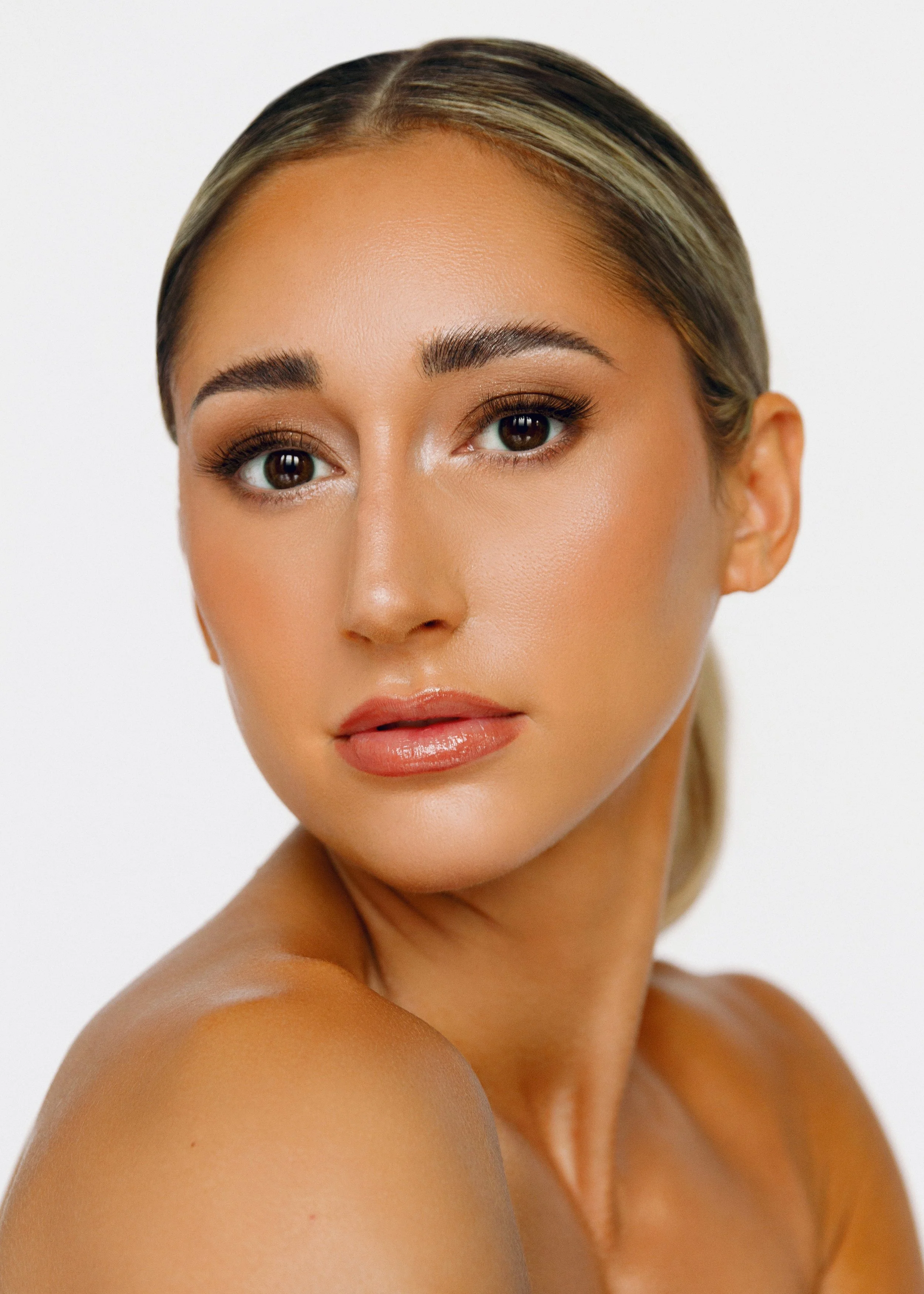 Close-up portrait of a woman with smooth, tan skin, dark eyebrows, subtle makeup, and long eyelashes. Her hair is blonde with darker roots, styled sleek and pulled back. She is looking slightly to the side with a calm expression. The background is plain white.