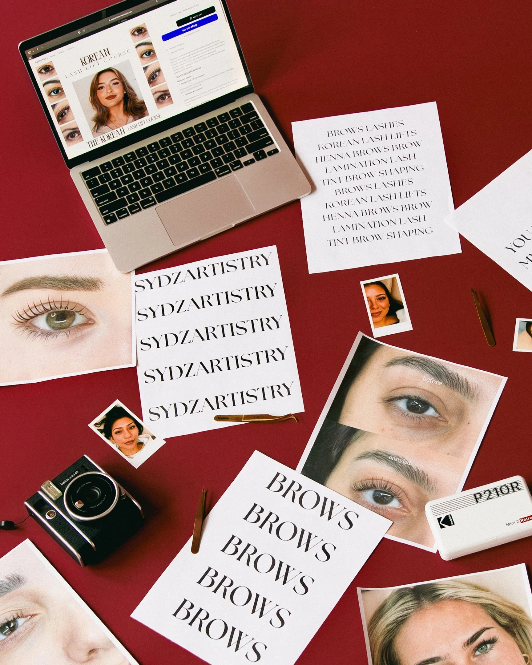 A laptop on a red table displaying a beauty course webpage. Surrounding papers with images of eyes, eyebrows, and makeup, and printed text including "BROW'S" and "SYDZARTISRY." There is a small camera and a pen on the table.
