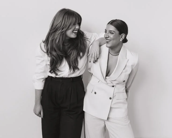 Two women smiling and laughing together, one with long wavy hair wearing a white blouse and high-waisted pants, the other with her hair in a bun wearing a white blazer and matching pants, standing against a plain background.
