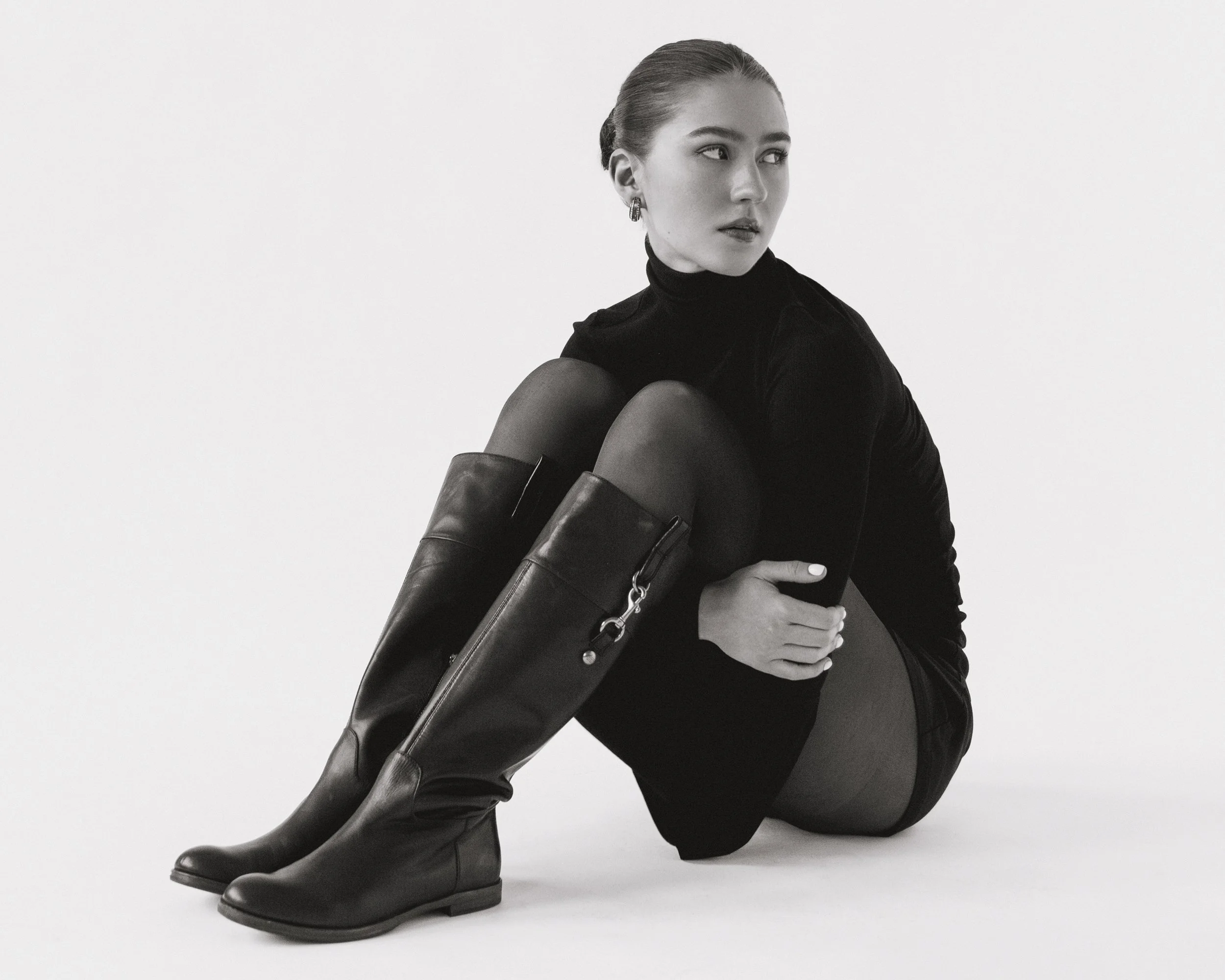 Black and white photo of a woman with her arms around her bent knees, sitting sideways on the floor, wearing a velvety black turtleneck, black leather thigh-high boots, and large earrings, against a plain white background.
