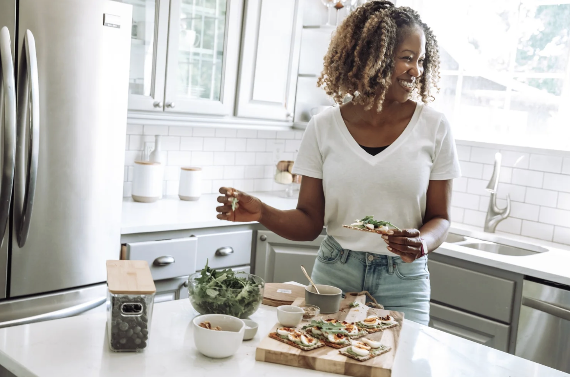 Smiling woman preparing healthy food in a bright kitchen, making fresh toast with greens for wellness, mindful eating, and balanced living.