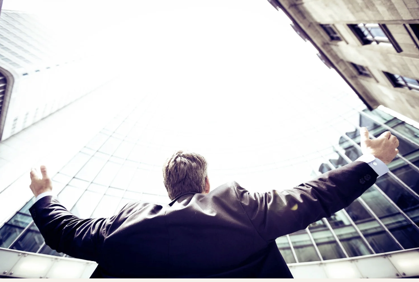 Businessman standing between tall city buildings with arms raised, symbolizing success, confidence, achievement, leadership, and personal growth.