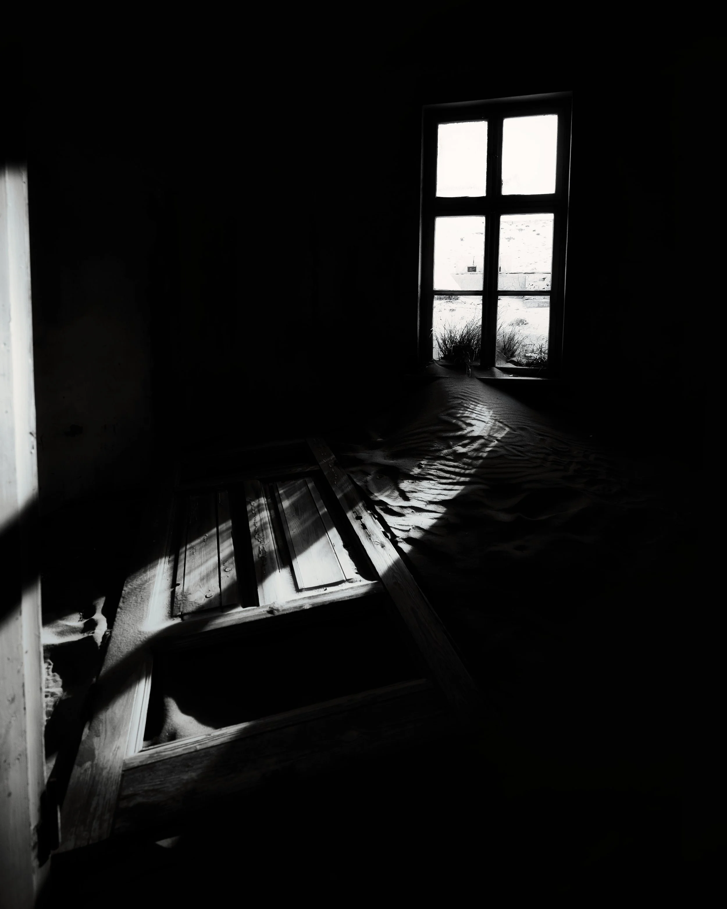 Dark room with a window letting in light, an overturned wooden door or frame on the floor, and a crumpled sheet or fabric.