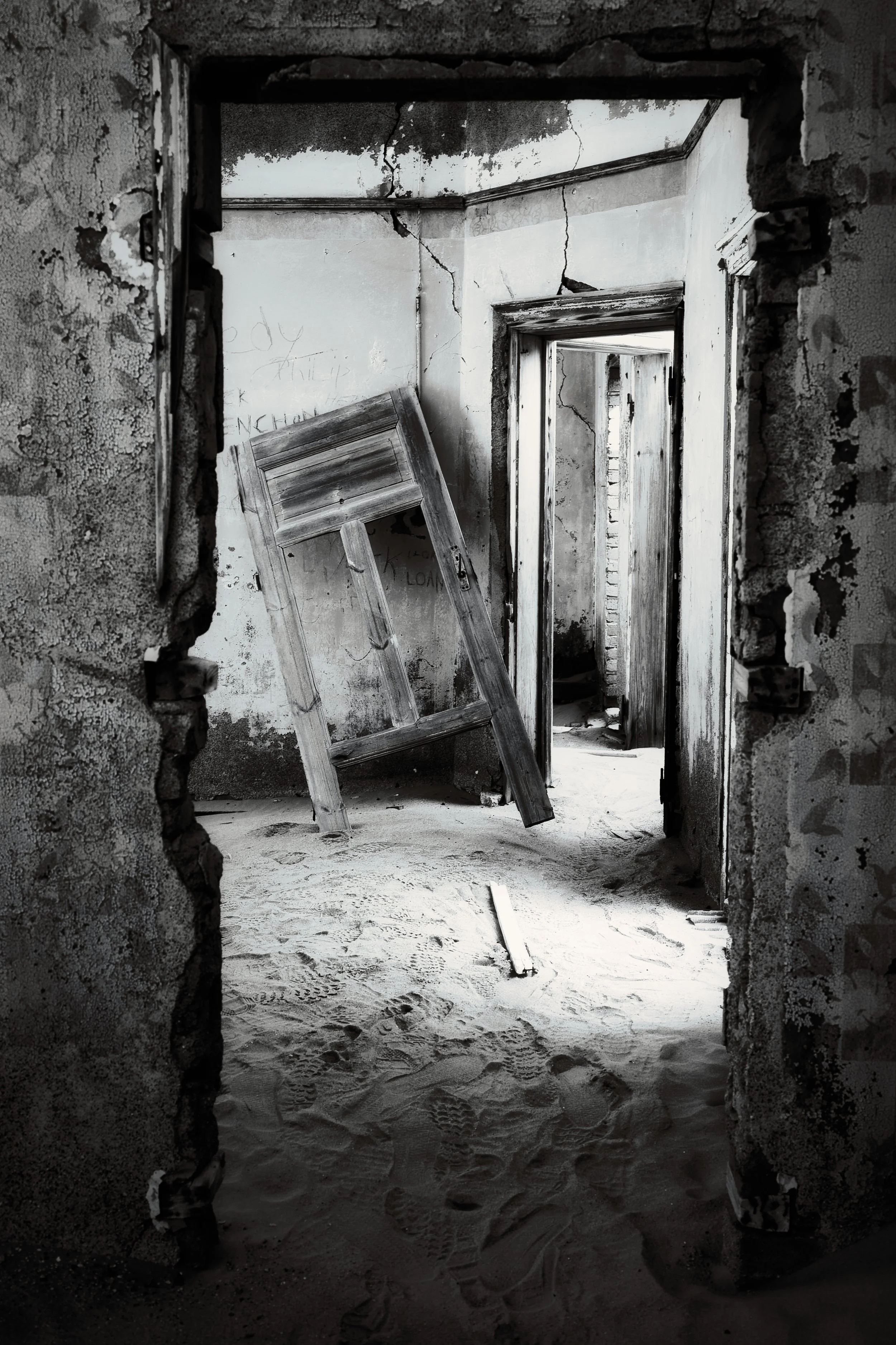 An abandoned, dilapidated building with cracks on the walls, a fallen wooden door, and sandy footprints on the floor.