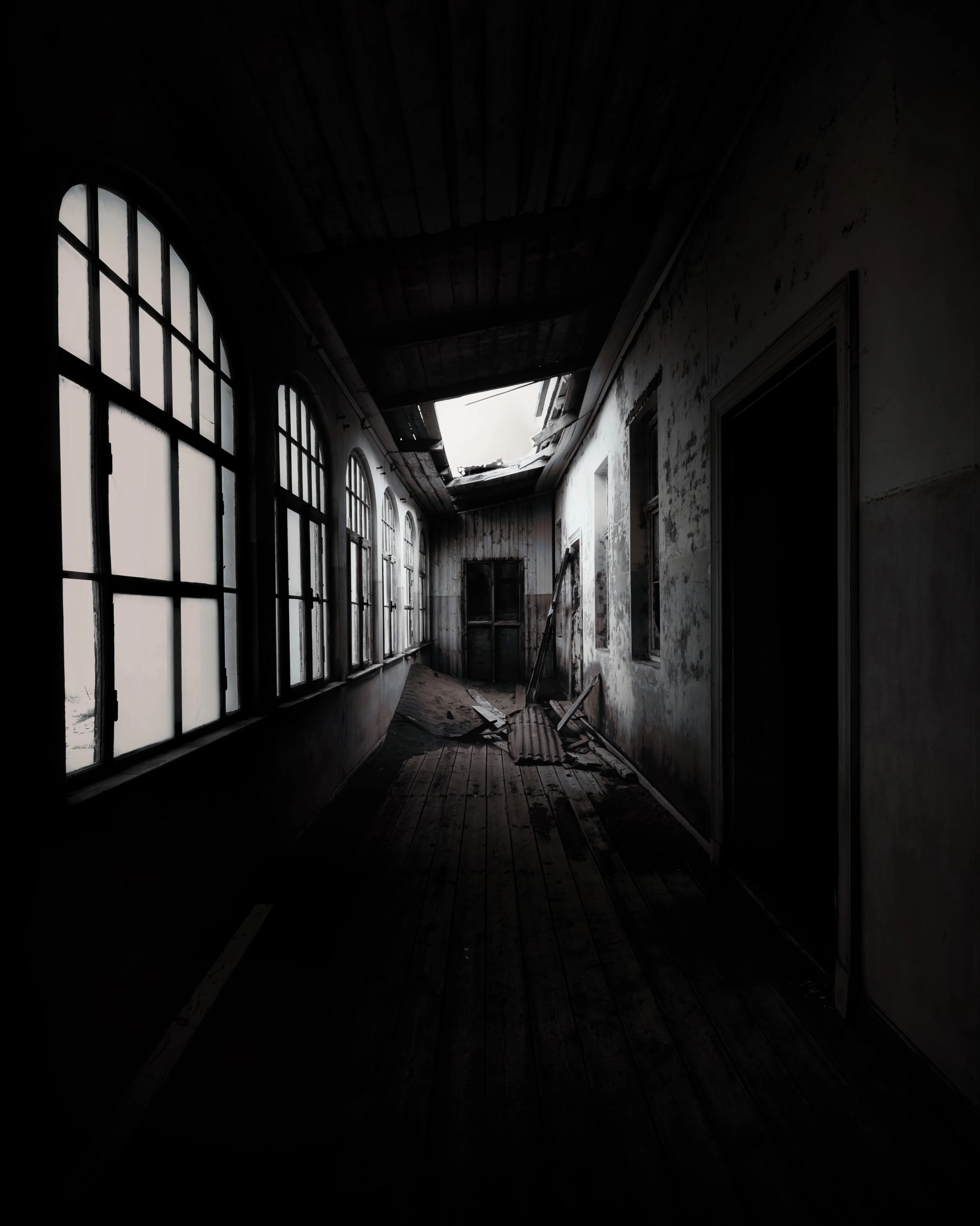 Dilapidated hallway with broken roof, shattered windows, debris on the floor, and peeling paint on the walls.