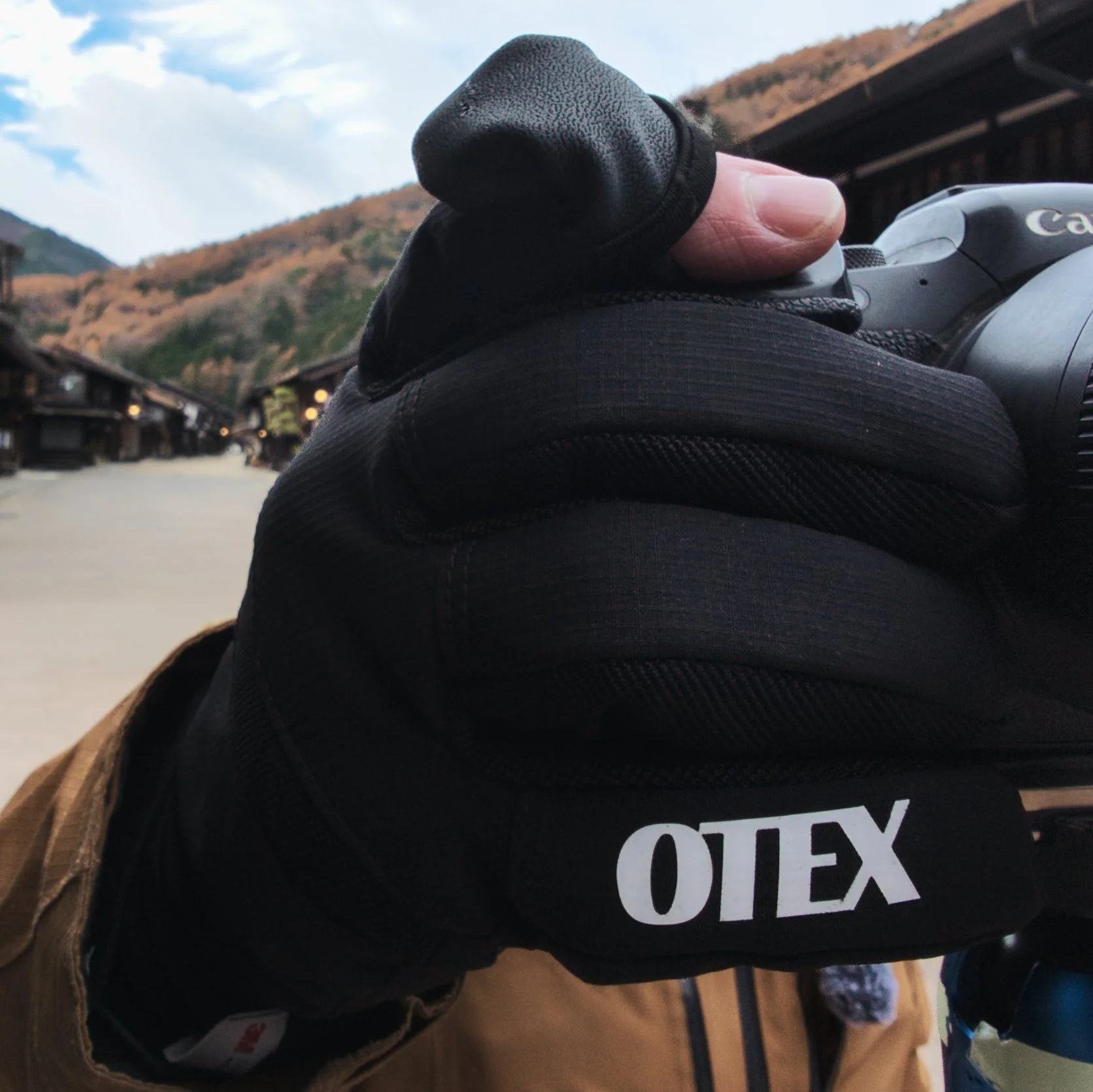 Close-up of a gloved hand holding a camera outdoors with a mountainous landscape and buildings in the background.