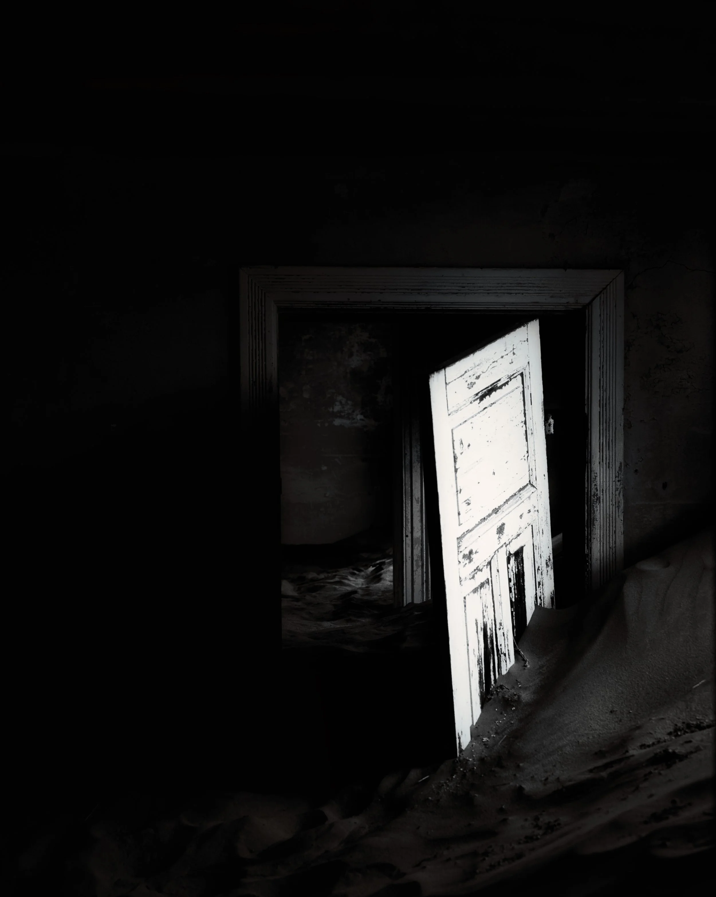 An old, white wooden door partially open, leaning and stuck in sand inside a dark, abandoned building.