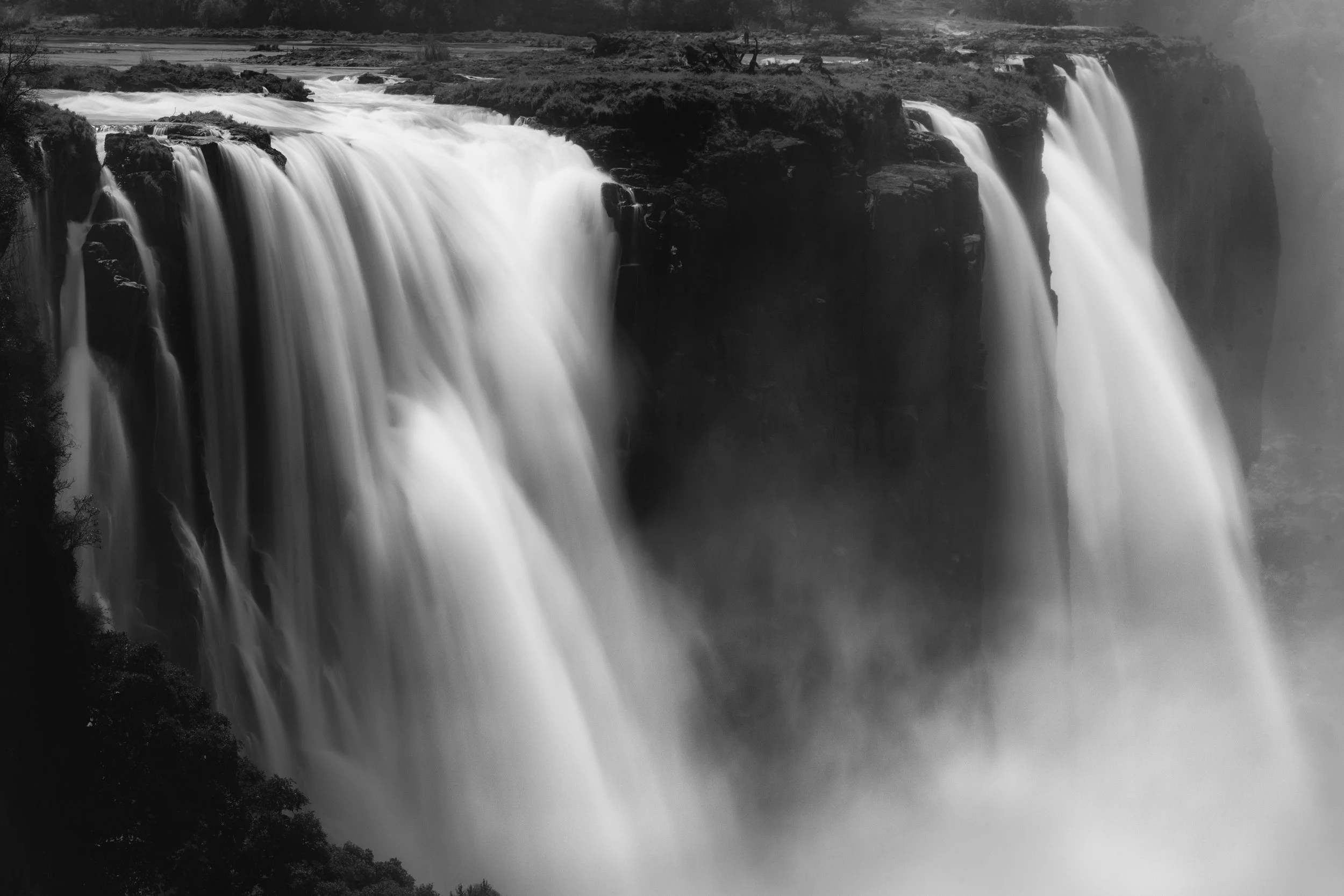 Black and white photo of a large waterfall with water cascading over cliffs into a misty pool below.