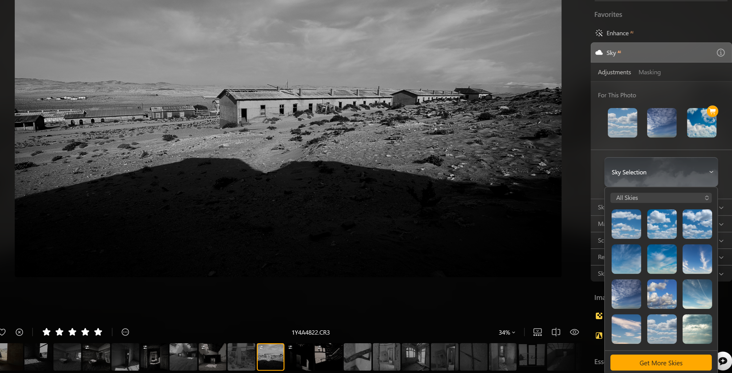 A black and white photo of an arid, rocky landscape with several old, abandoned buildings in the background under a partly cloudy sky.