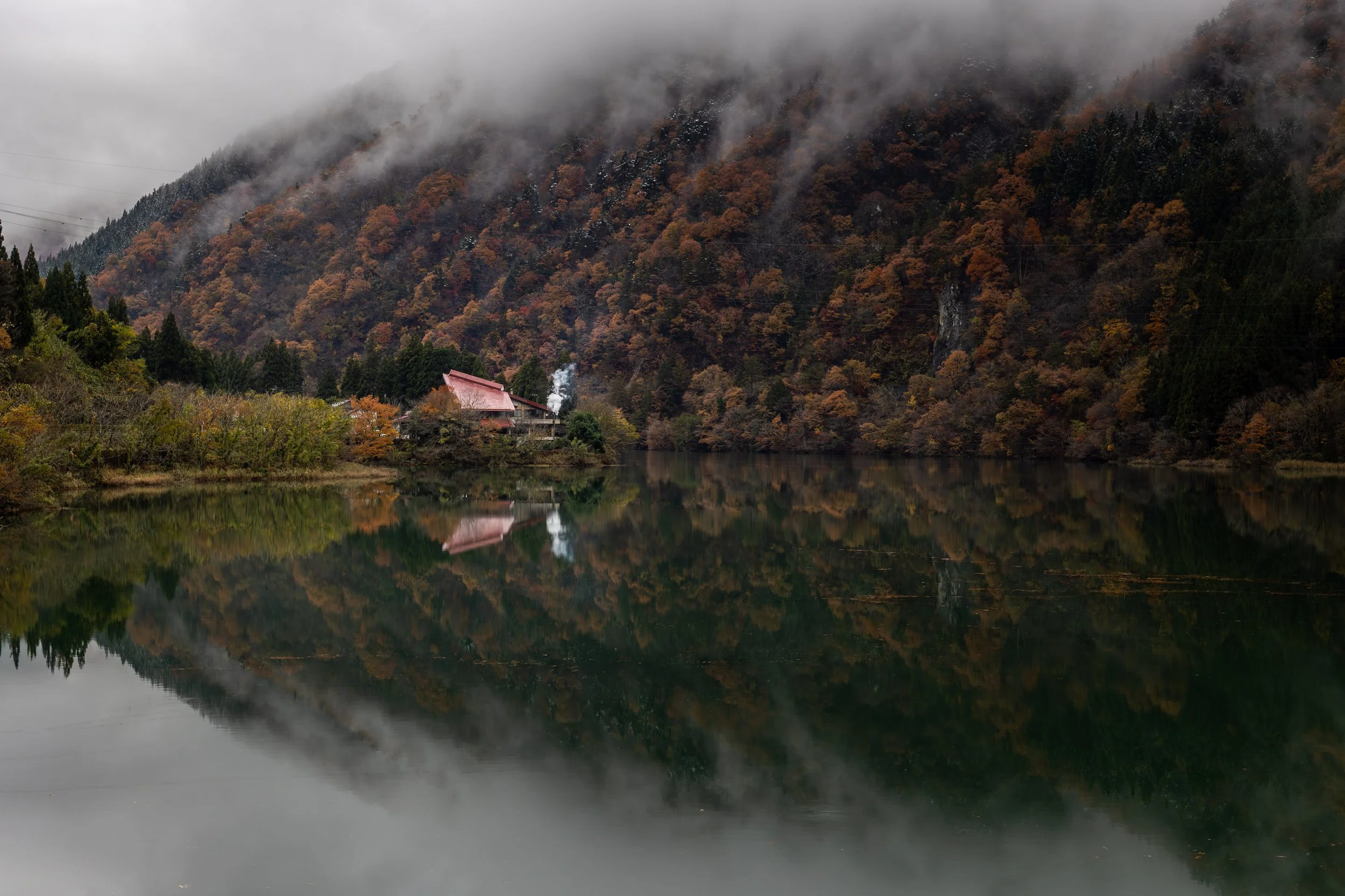 A serene lake reflecting a dense, colorful autumn forest and mountains shrouded in mist, with a small house with a red roof and smoke rising from its chimney.