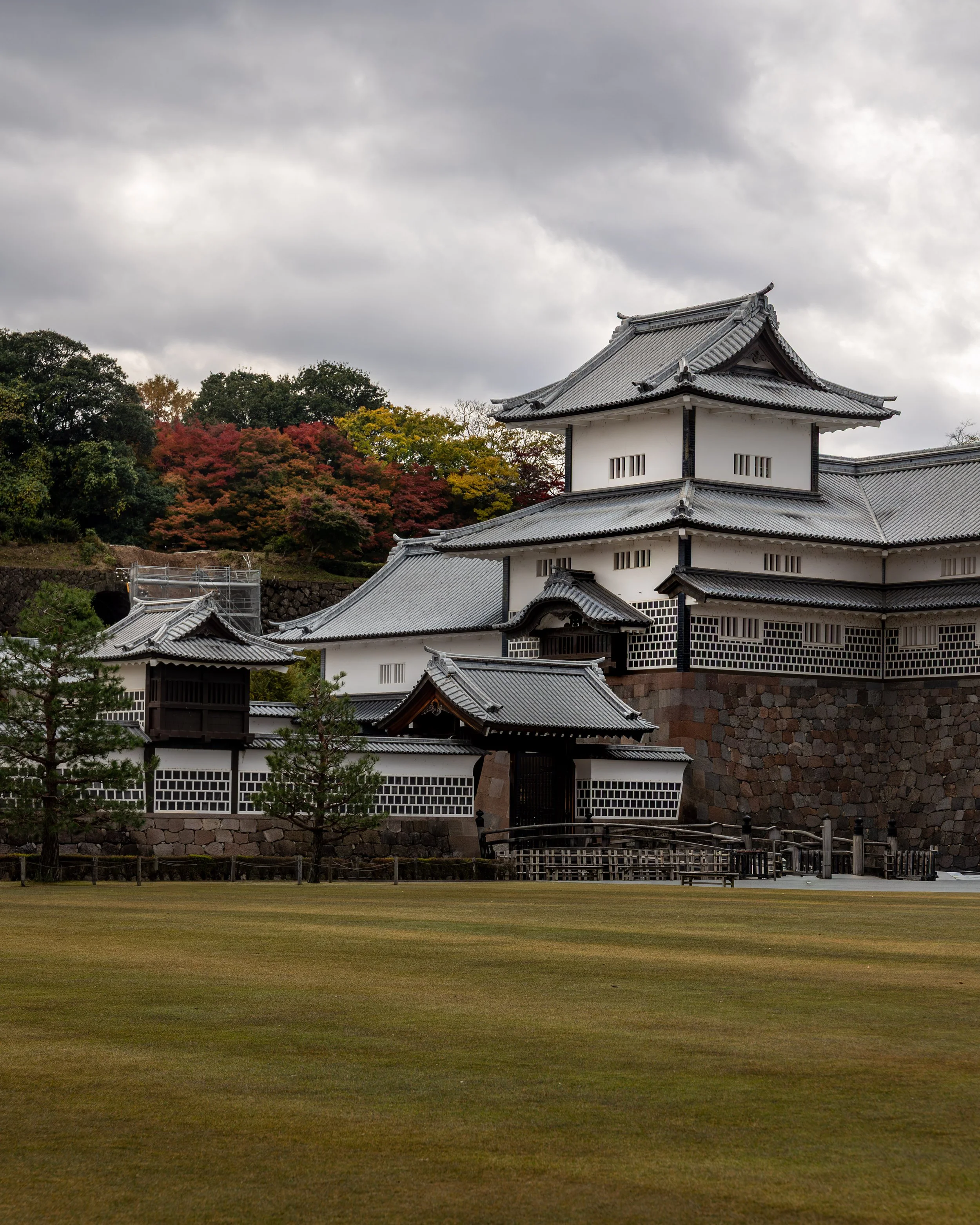 A traditional Japanese castle with white walls, gray tiled roofs, surrounded by trees with autumn foliage, and a grassy foreground under an overcast sky.