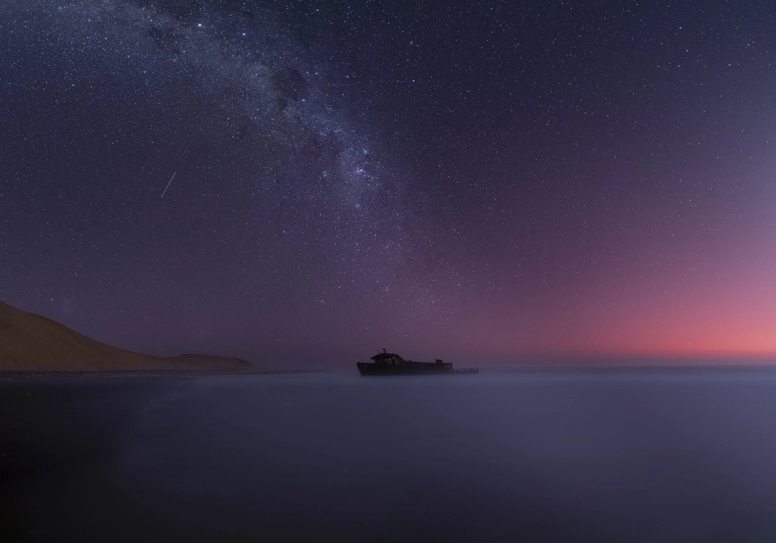 Nighttime scene of the ocean with a boat silhouetted against a colorful horizon, starry sky and the Milky Way galaxy visible overhead.