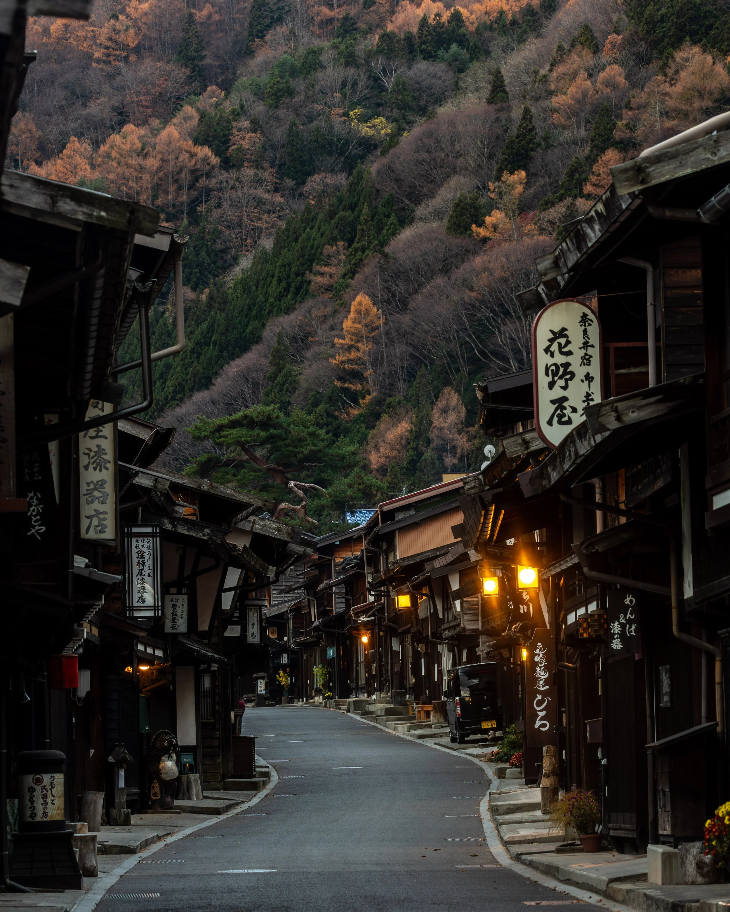A quiet street in a traditional Japanese village surrounded by a hillside with colorful autumn trees, bamboo, and evergreens, with wooden buildings and glowing lanterns.