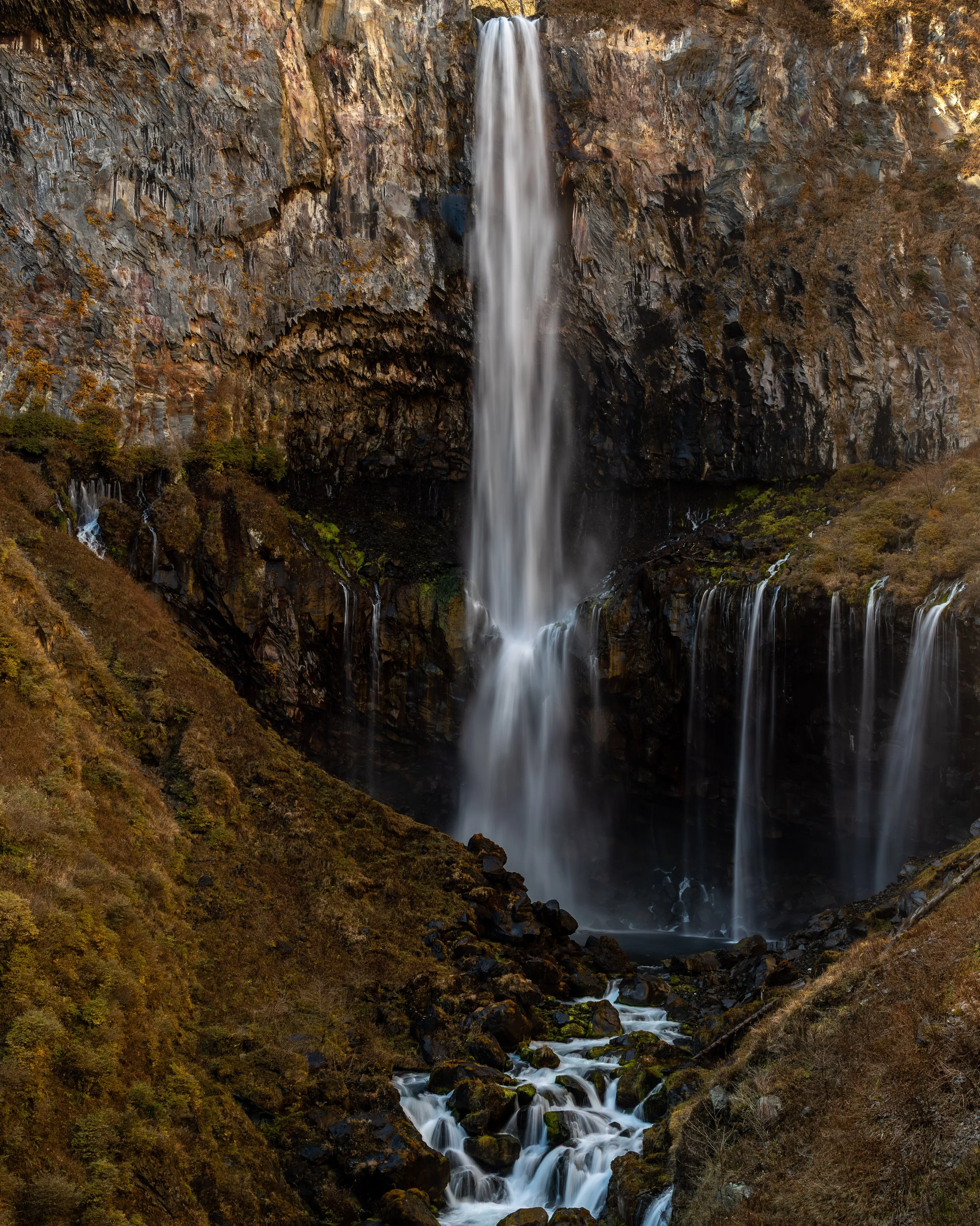 A tall waterfall cascading down a steep, rocky cliff surrounded by foliage and mossy rocks.