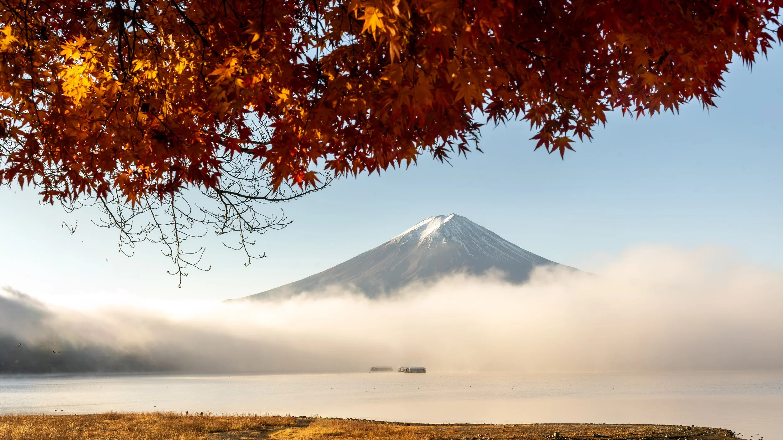 Autumn leaves in orange and red in the foreground with snow-capped Mount Fuji in the background and a lake with a partially cloudy sky.