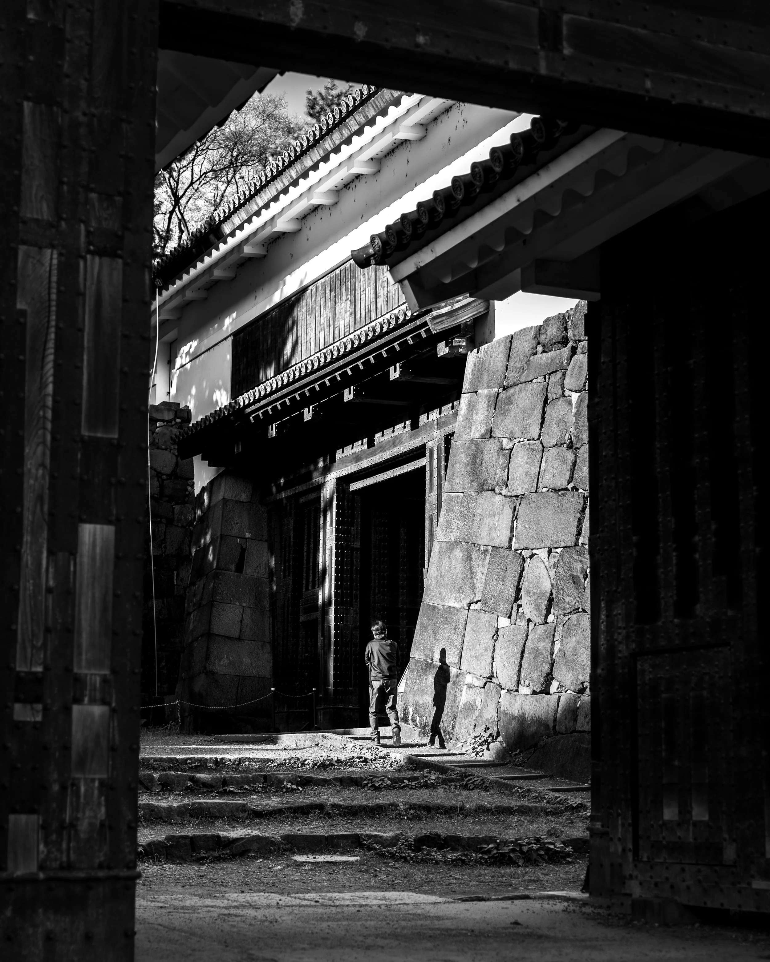 A person walking through a traditional Japanese temple gate with stone steps and walls, black and white photograph.