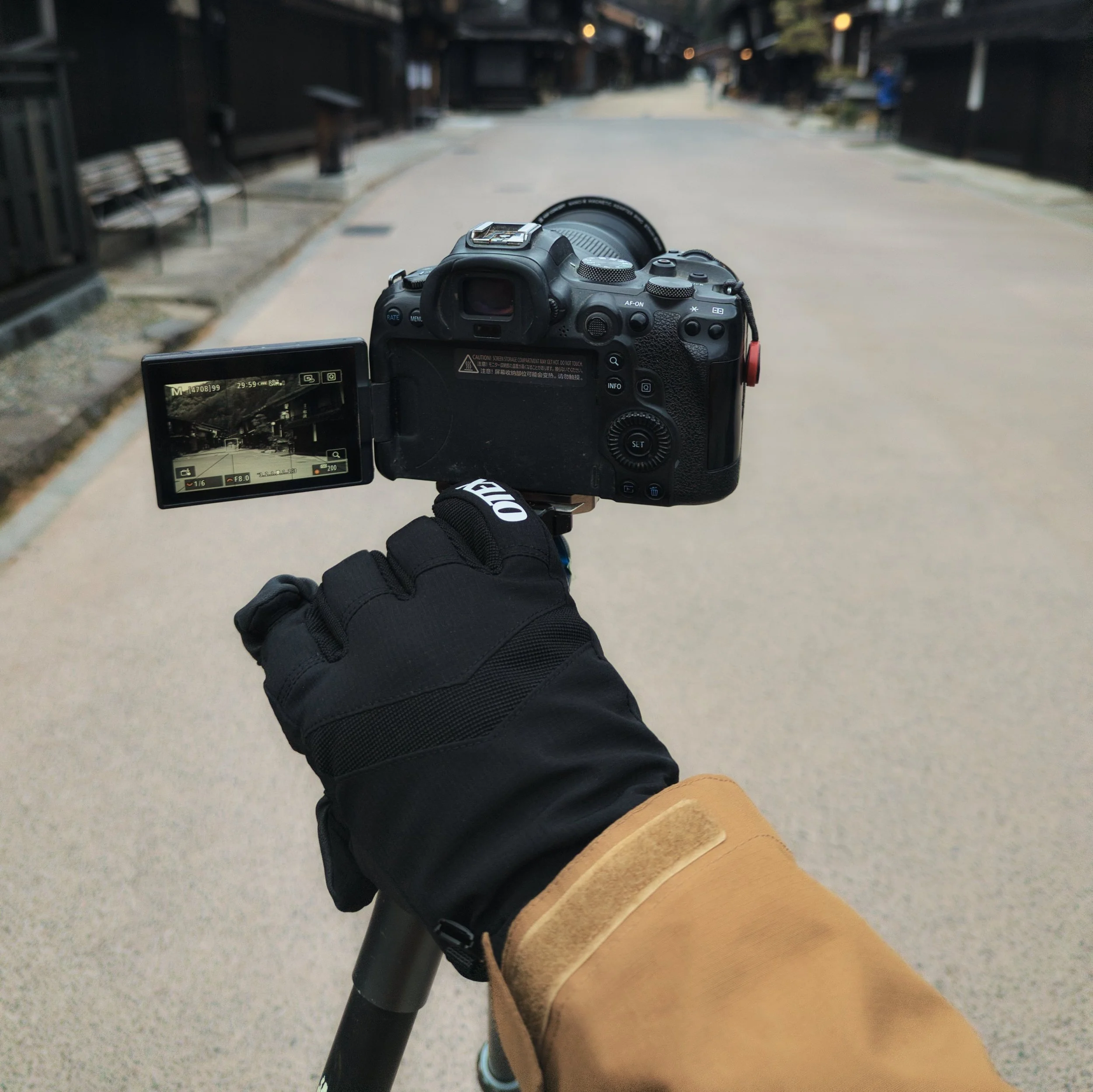 A person wearing a black glove and tan coat holding a camera on a tripod, aimed down an empty, paved street lined with black buildings, benches, and lampposts.
