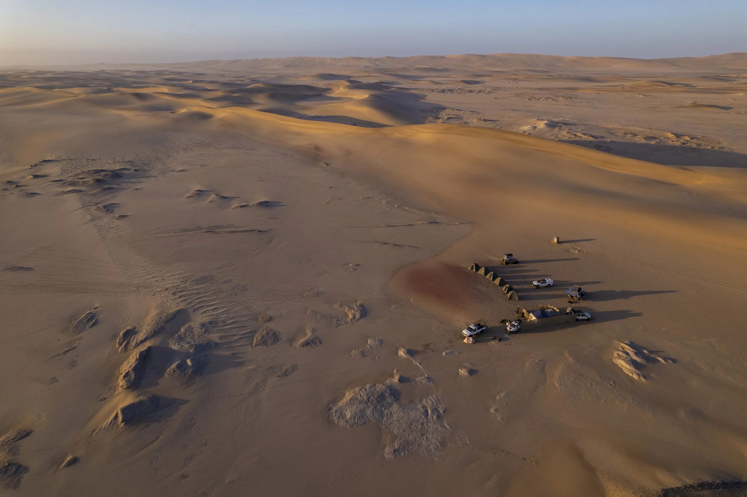 A desert landscape with sand dunes and a small encampment of vehicles and tents.