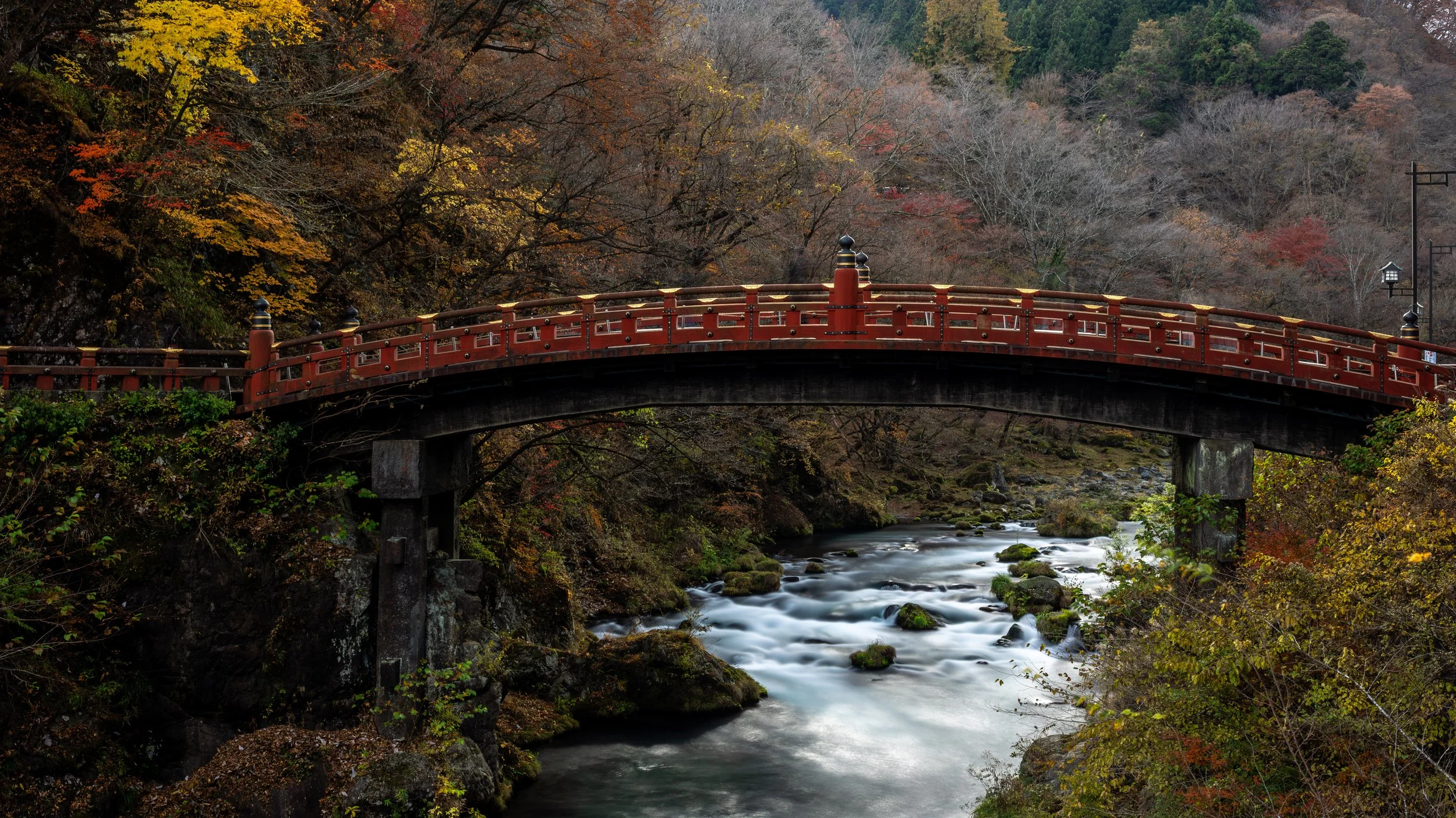 A red arched bridge over a flowing river surrounded by autumn trees with colorful leaves.