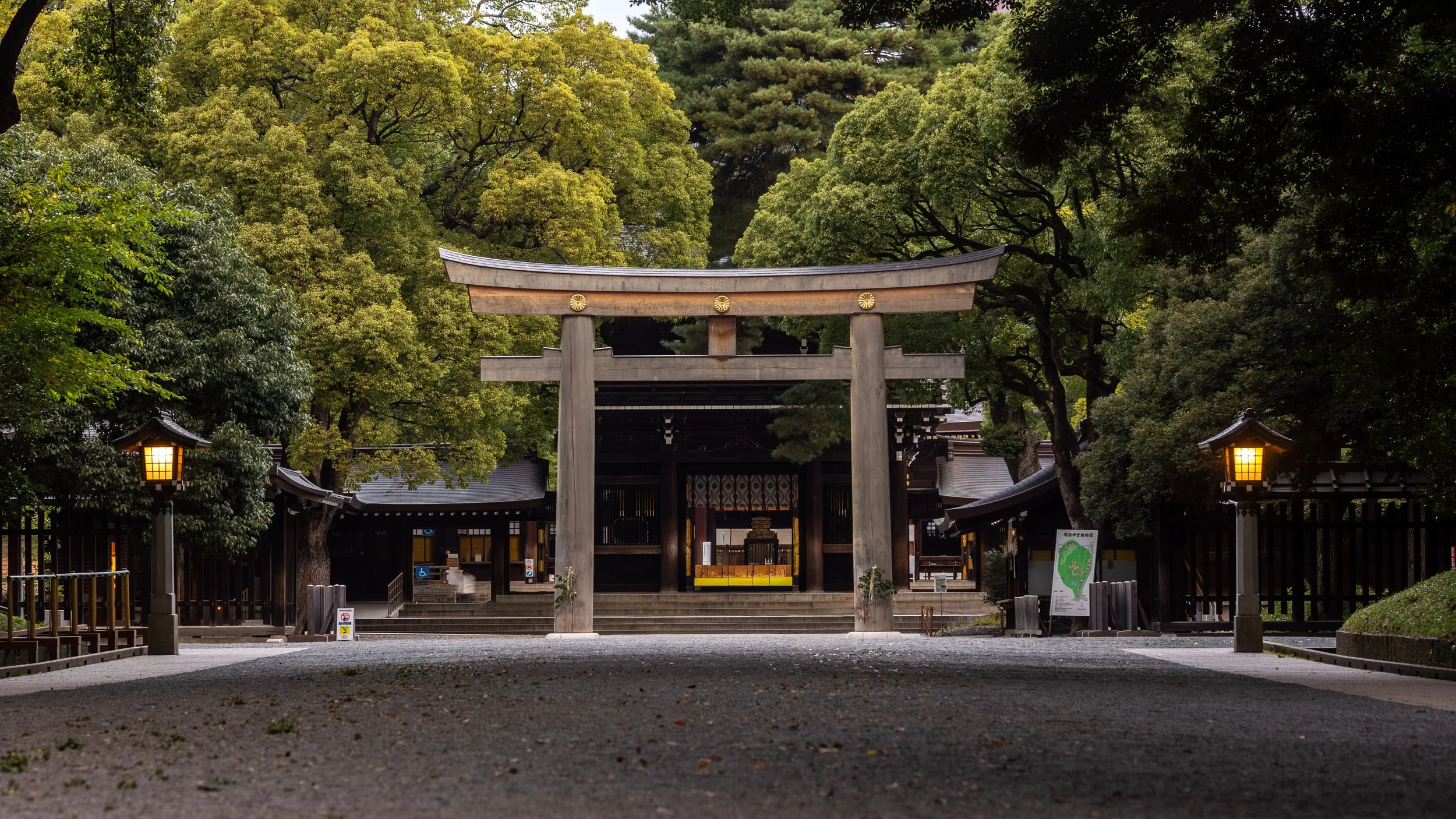A traditional Japanese shrine entrance with a large wooden torii gate, surrounded by green trees and lit lanterns on either side.
