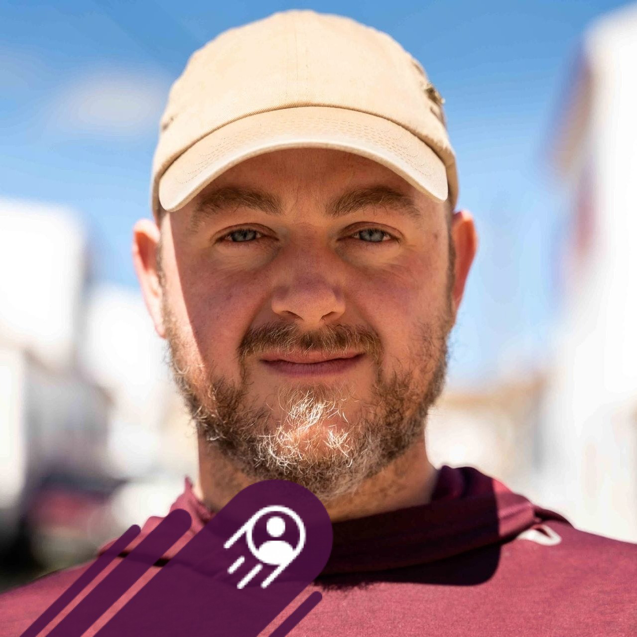 Close-up of a man with a beard and mustache wearing a beige baseball cap and maroon shirt, smiling outdoors on a sunny day, with blurred background and a purple icon with a white person and motion lines overlayed at the bottom.