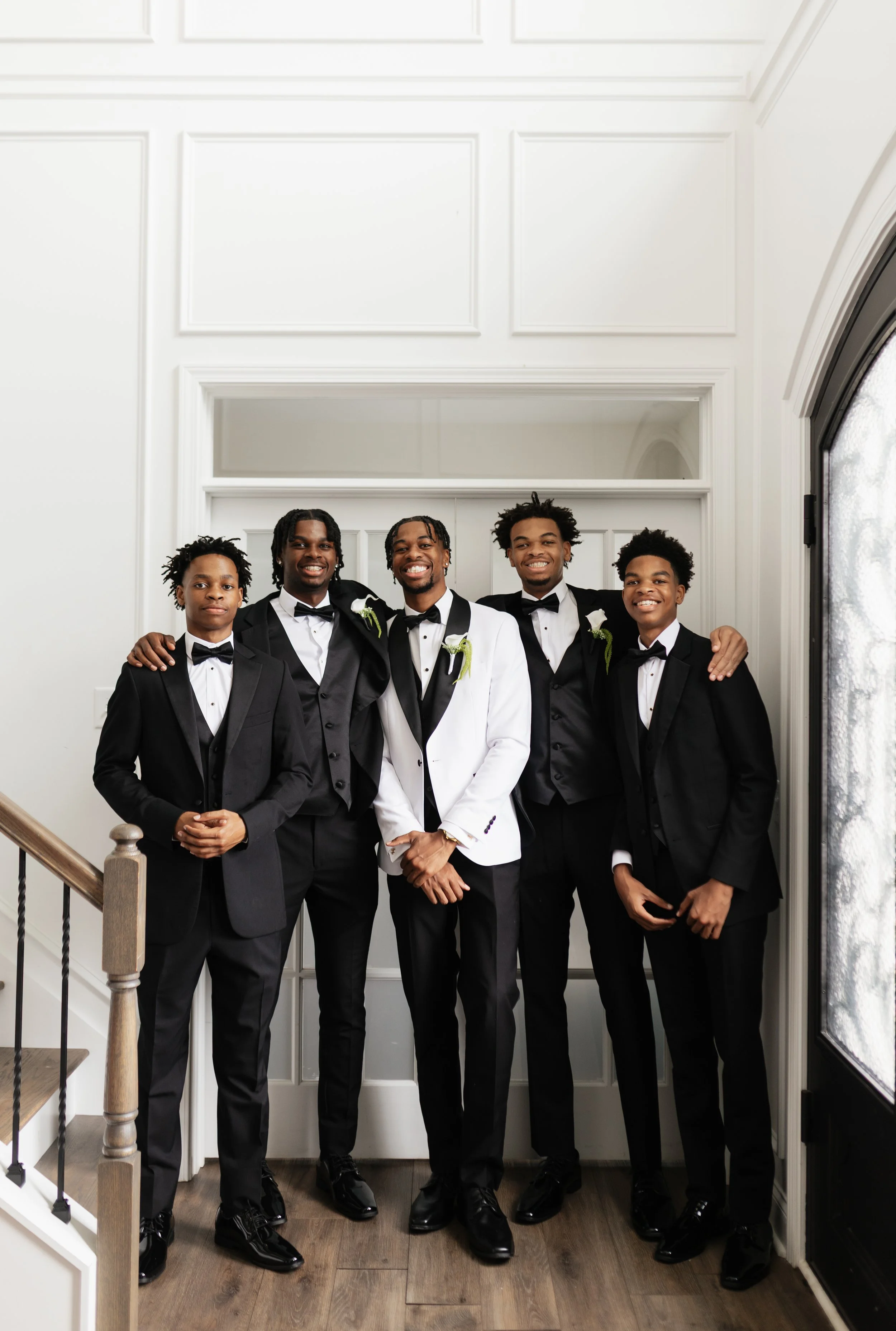 Group of five young men dressed in tuxedos and bow ties, standing indoors near a white paneled wall, smiling for a photo.