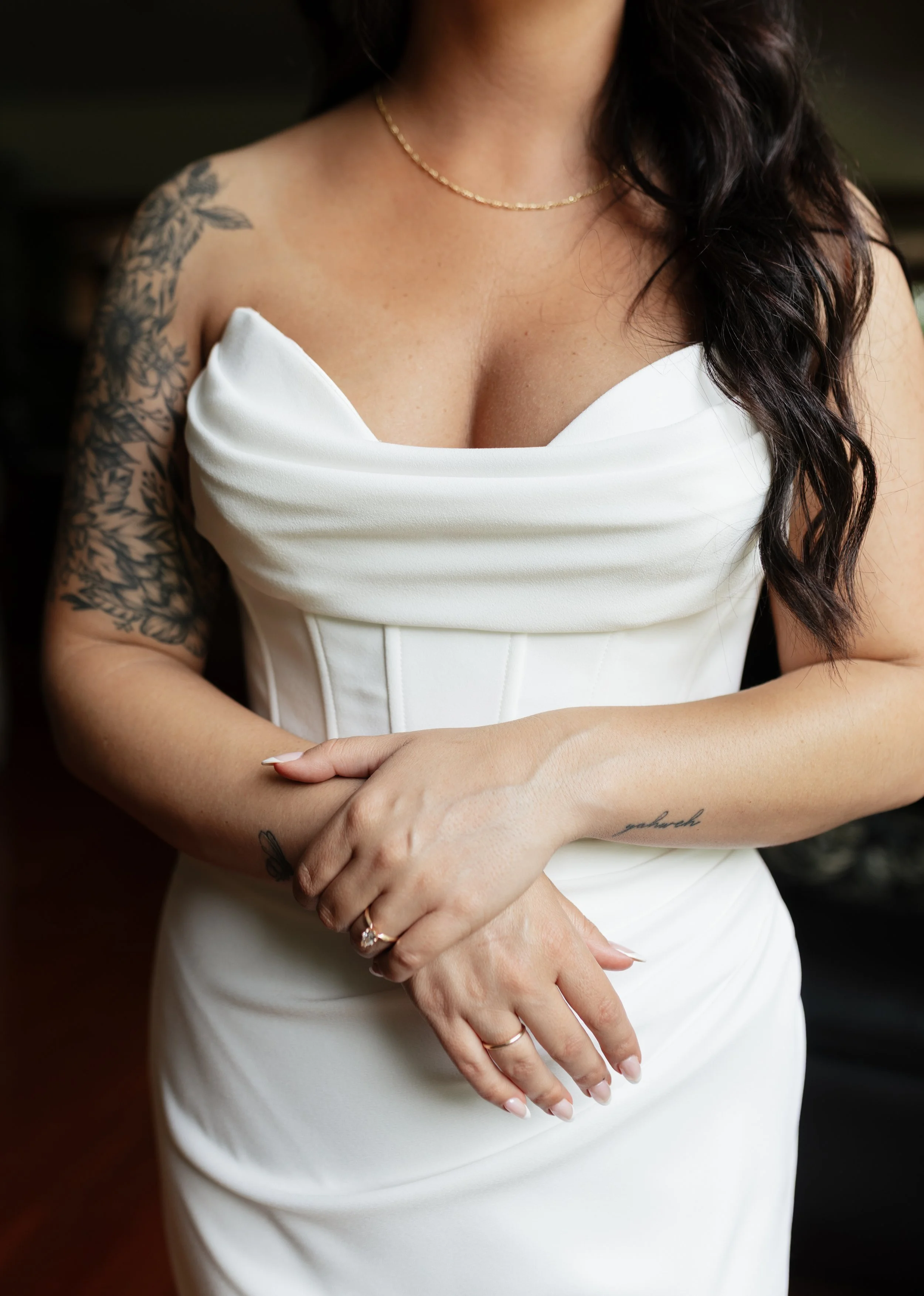 Close-up of a woman wearing a white strapless dress, with long dark hair, wearing a thin gold necklace, with tattoos on her left arm, and with her hands crossed, showing a gold ring and a tattoo on her wrist.