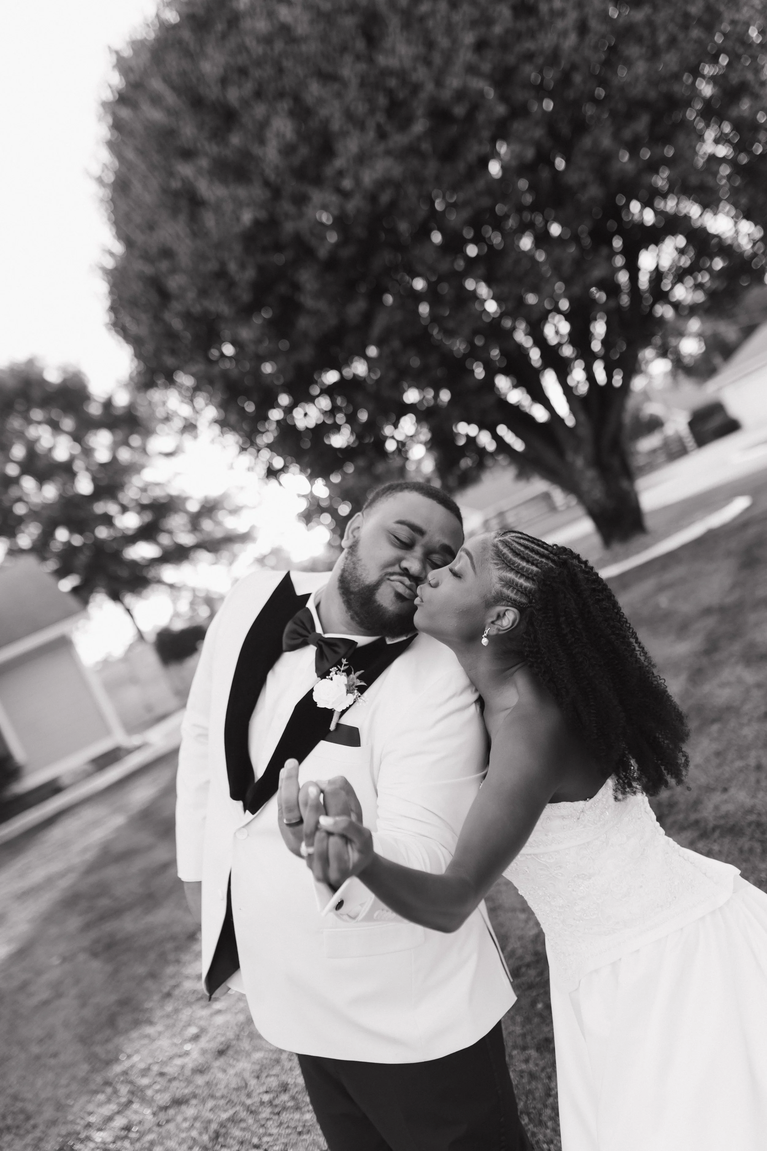 A black and white photo of a newlywed couple sharing a kiss outdoors. The groom is wearing a tuxedo with a bow tie, and the bride is in a wedding dress with braided hair. They are holding hands, and large trees are visible in the background.
