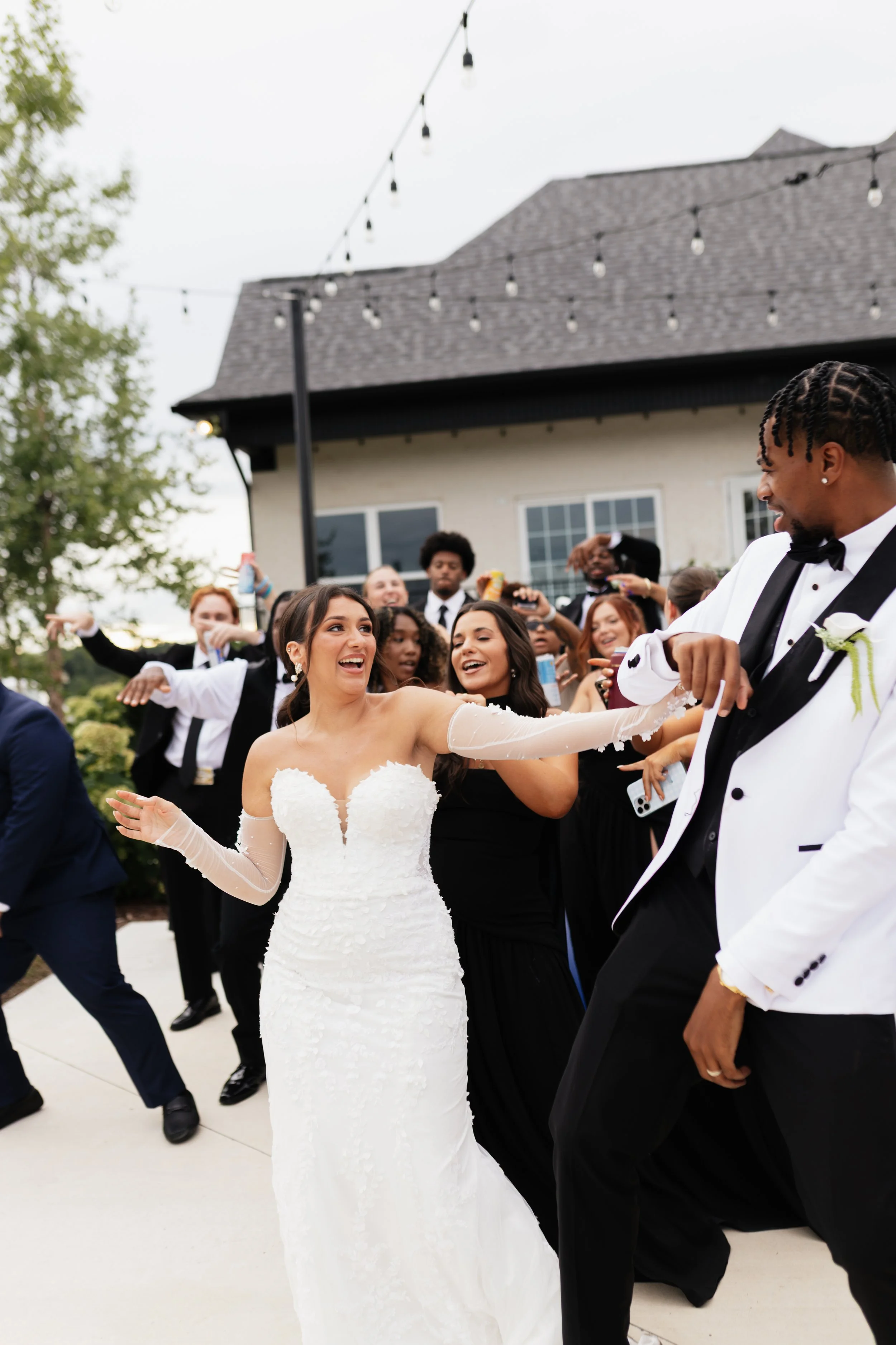 Wedding reception outdoors with guests dancing, including a bride in a white gown and a groom in a tuxedo, with string lights above and a house in the background.