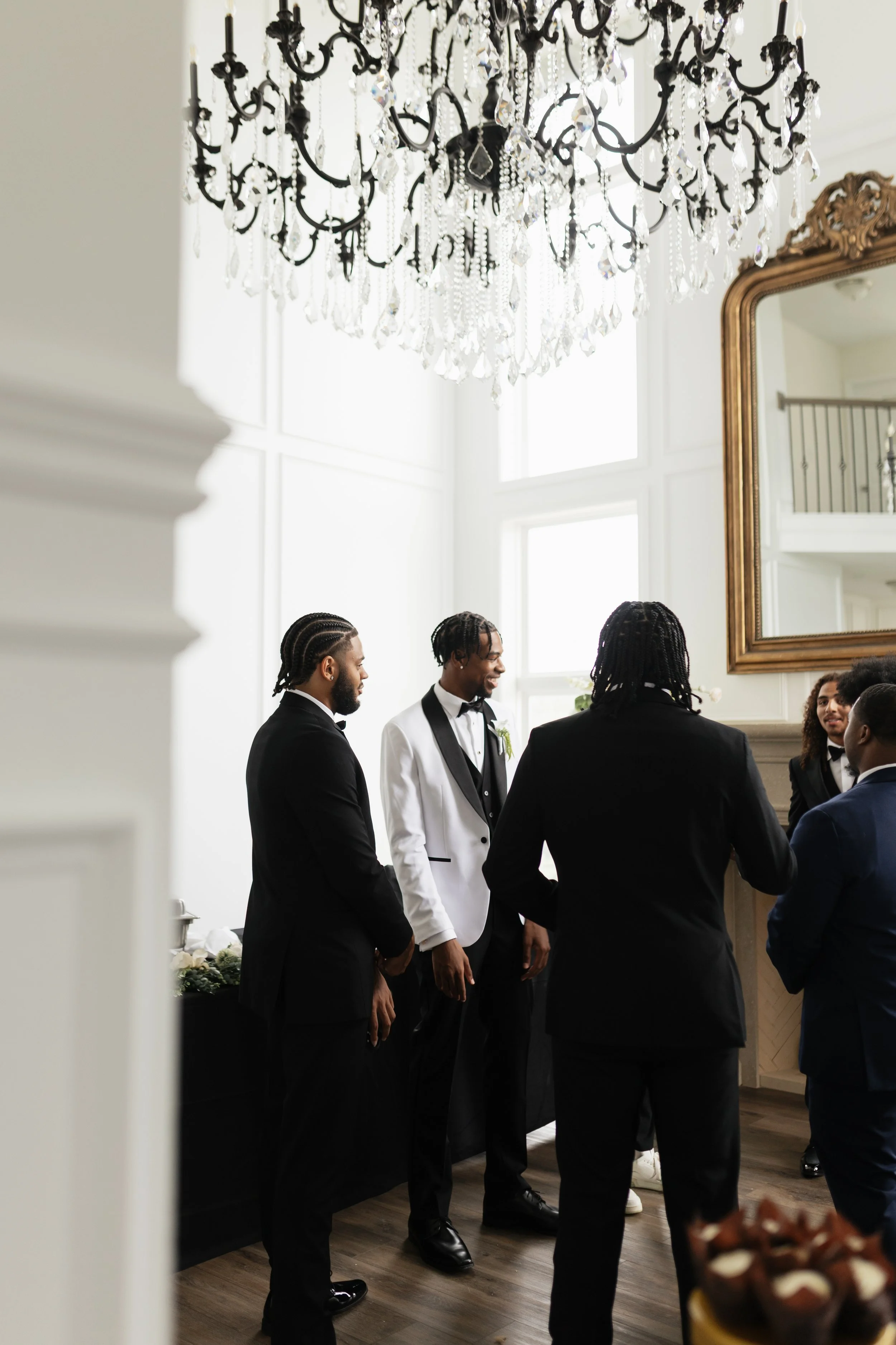 A group of men dressed in tuxedos having a conversation in a well-lit room with a large chandelier and a gold-framed mirror.