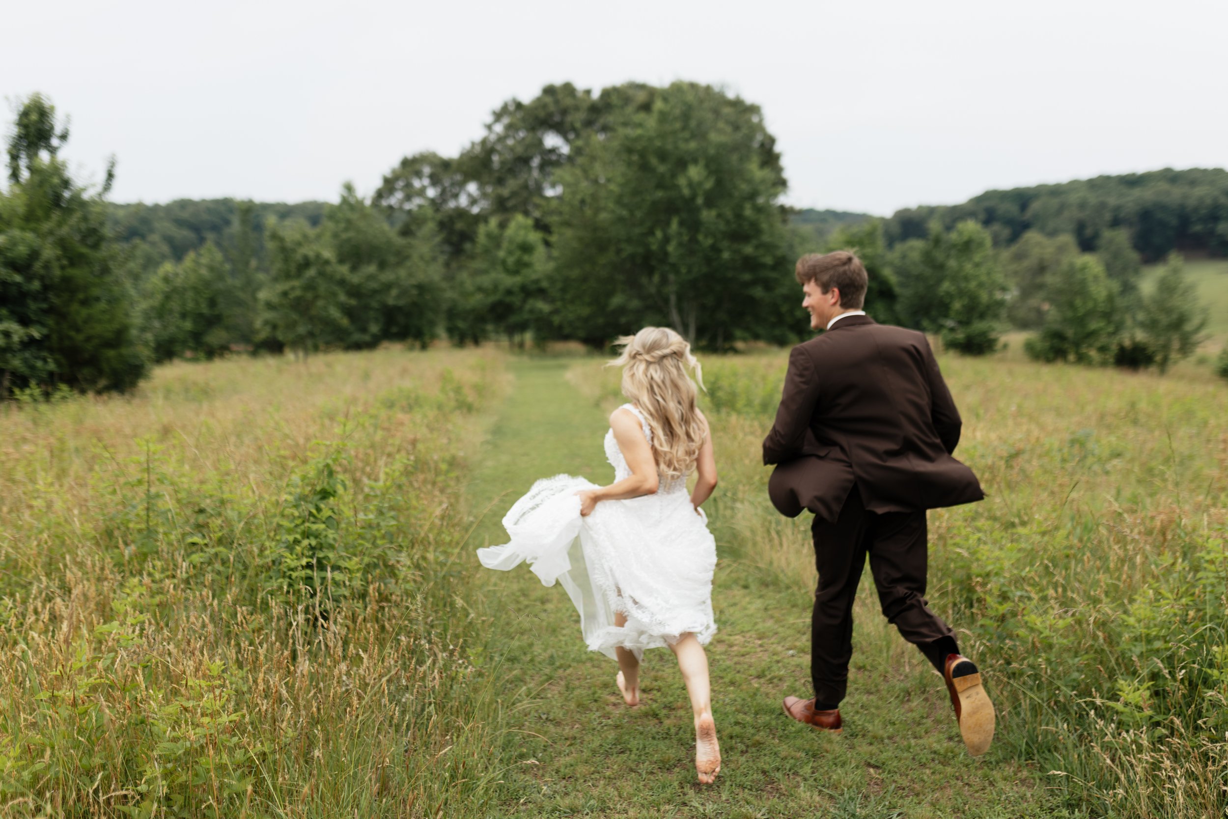 A couple running through a grassy field, with the woman wearing a white dress and the man dressed in a brown suit, surrounded by trees in a rural setting.