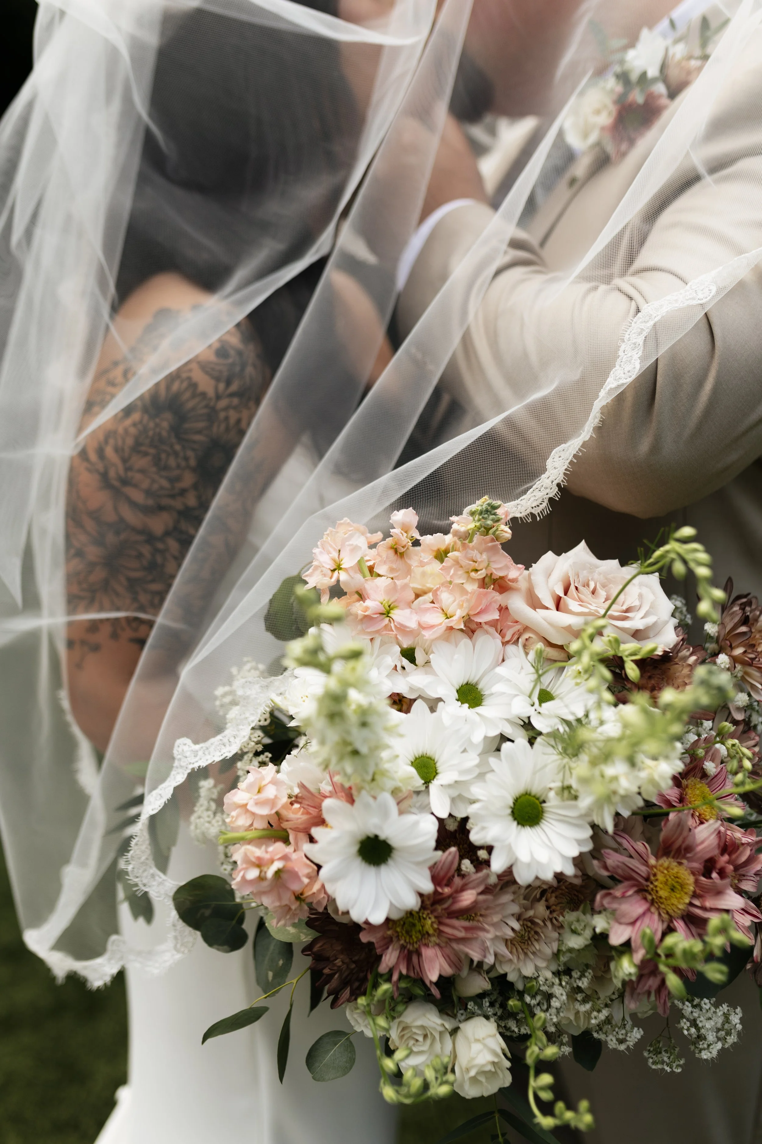 Close-up view of a wedding bouquet with white daisies, pale pink roses, and other flowers, wrapped in a veil with lace trim.